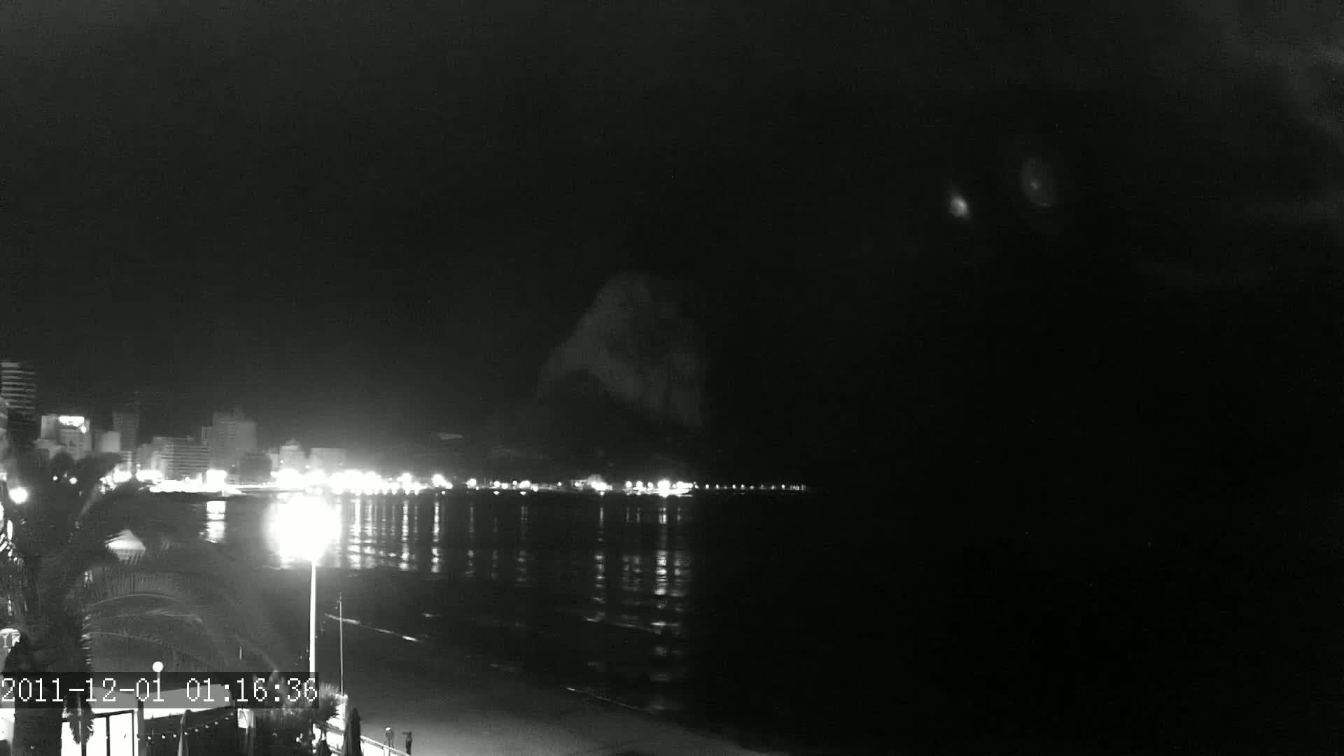 A nighttime coastal scene shows a dark, calm body of water reflecting the lights of a distant city, with a large, dark landmass rising behind it under a clear, dark sky.
