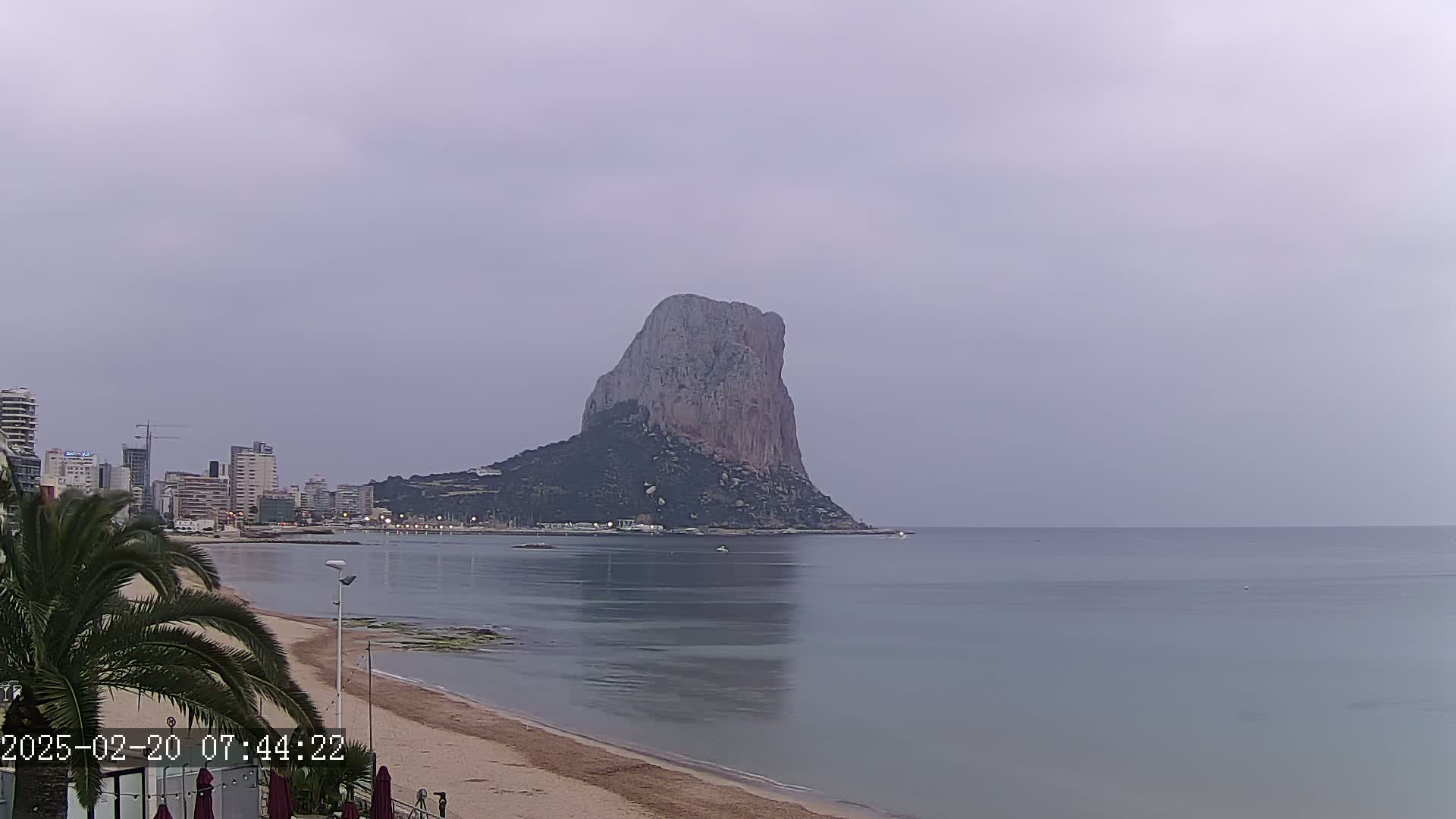 A large, dark rock formation rises from the water near a sandy beach and cityscape under an overcast sky.