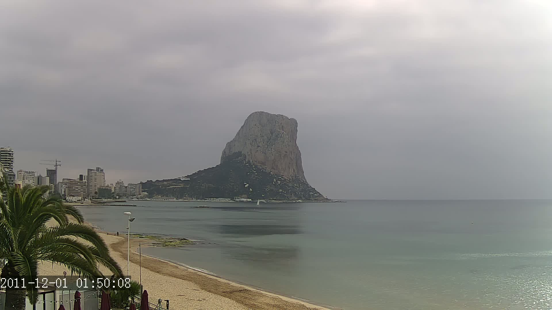 A large, rocky island rises from the calm, gray-green sea under an overcast sky, with a sandy beach and cityscape visible in the foreground.