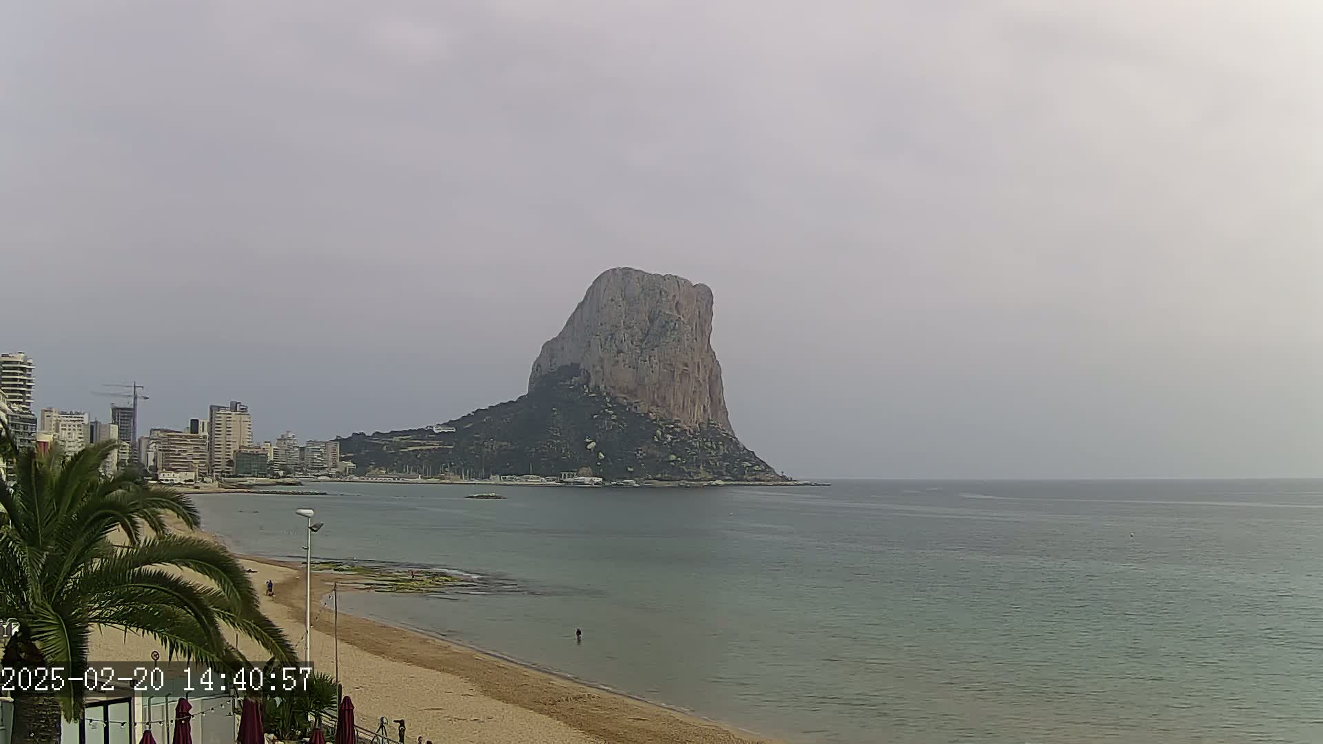 A large, rocky island sits in a calm, gray-toned sea off a sandy beach with buildings and palm trees in the foreground under an overcast sky.