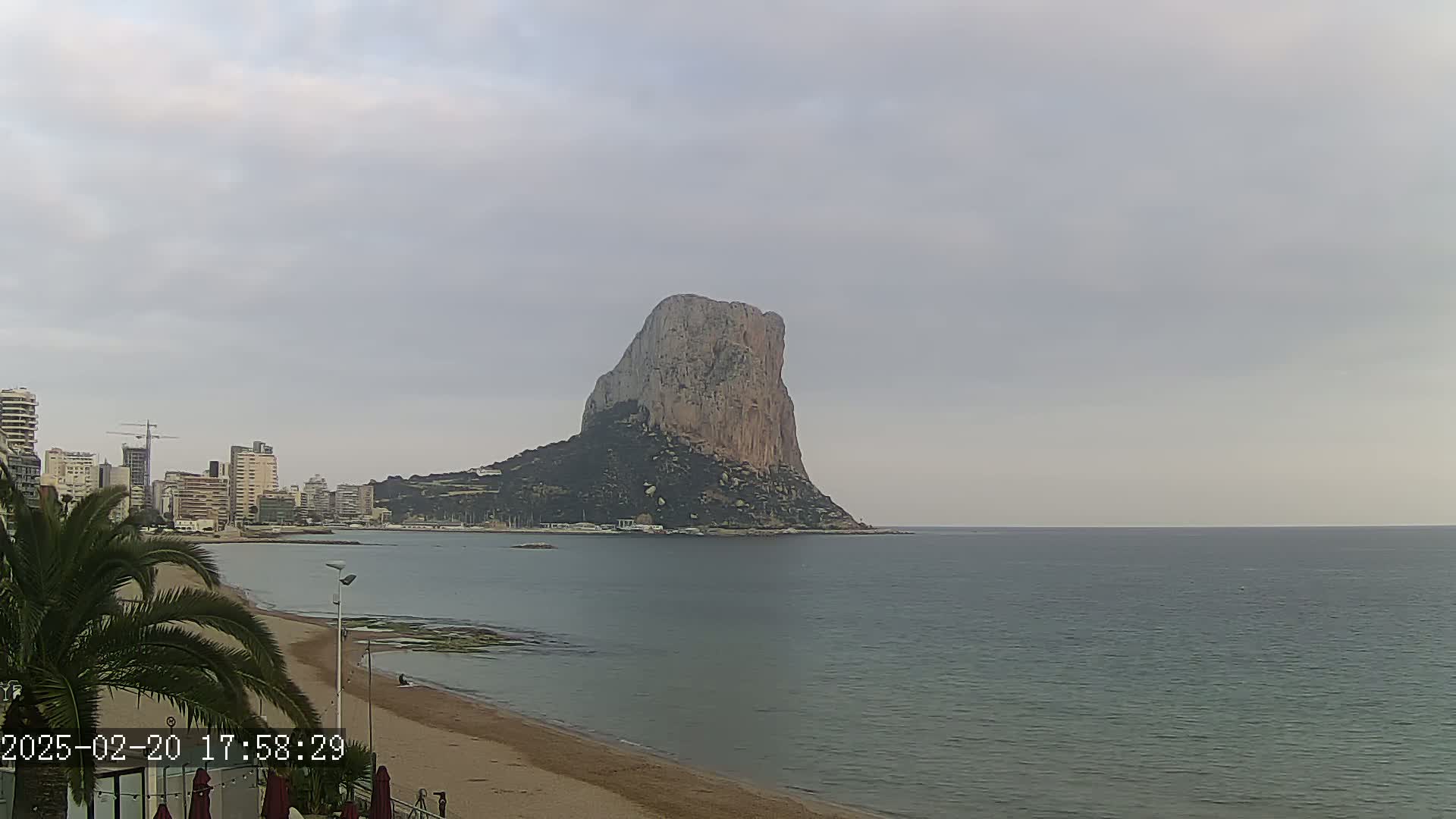 A large, rocky island sits in the calm ocean under an overcast sky, with a sandy beach and cityscape visible in the foreground.