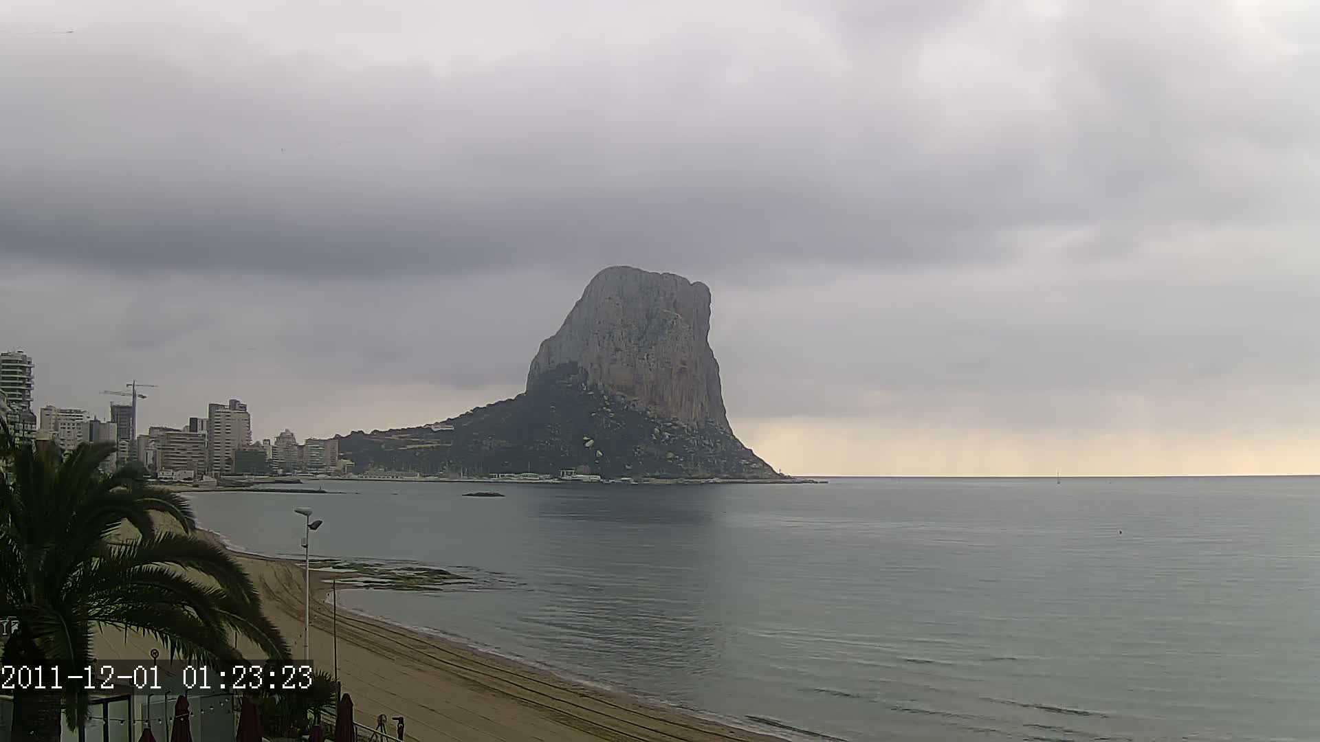 A large, solitary rock formation rises from the sea under an overcast sky, with a city skyline and sandy beach visible in the foreground.