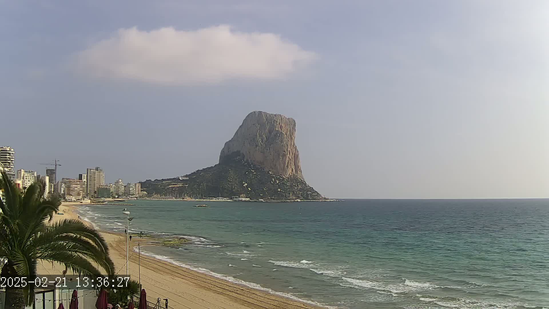 A large rock formation rises from the sea near a sandy beach and a city skyline under a partly cloudy sky.