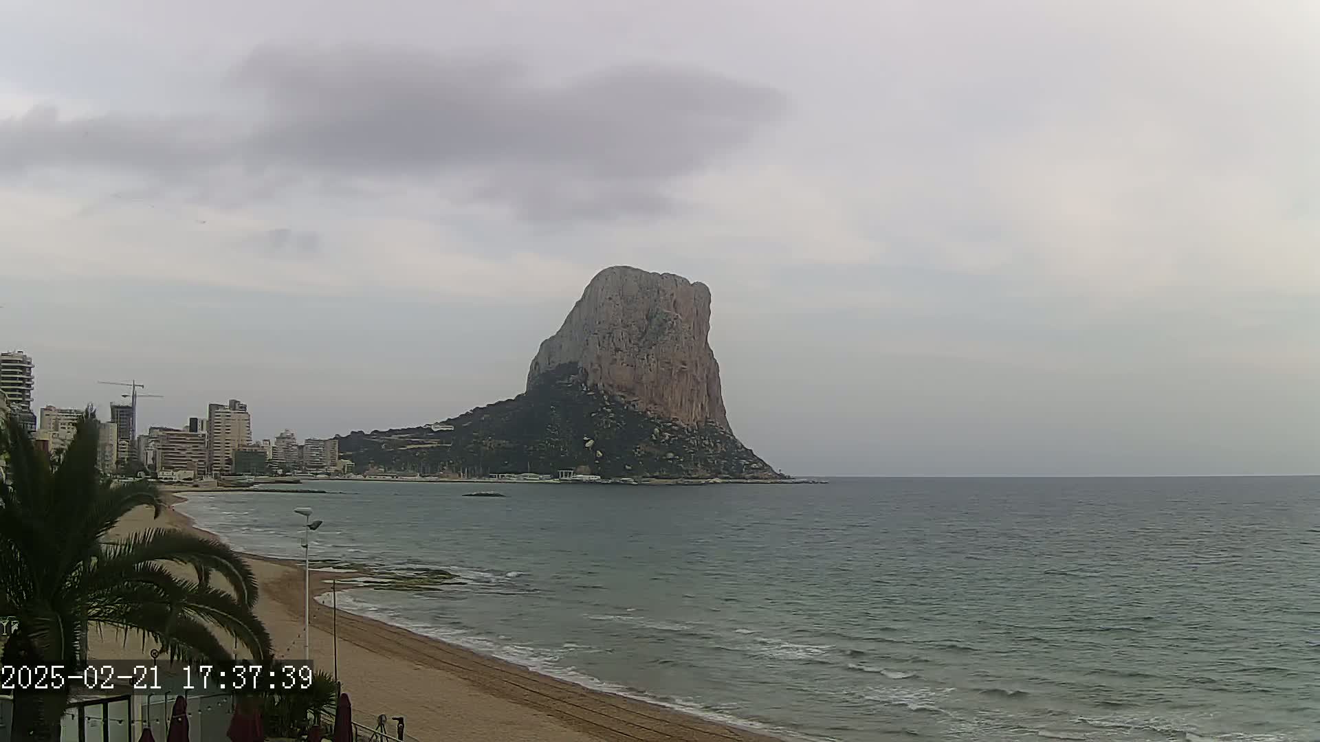 A large, rocky outcrop rises from the sea near a sandy beach and a city skyline under an overcast sky.