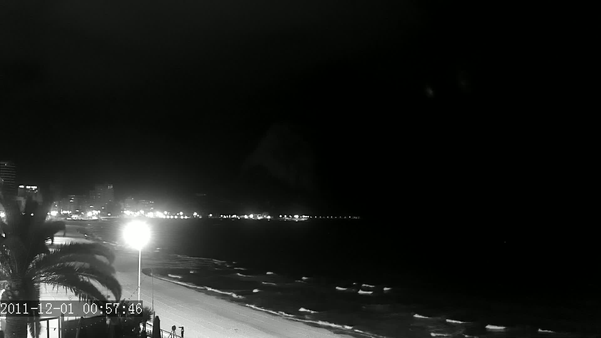 A nighttime beach scene shows gently lapping waves on a sandy shore, illuminated by city lights in the distance under a dark sky.