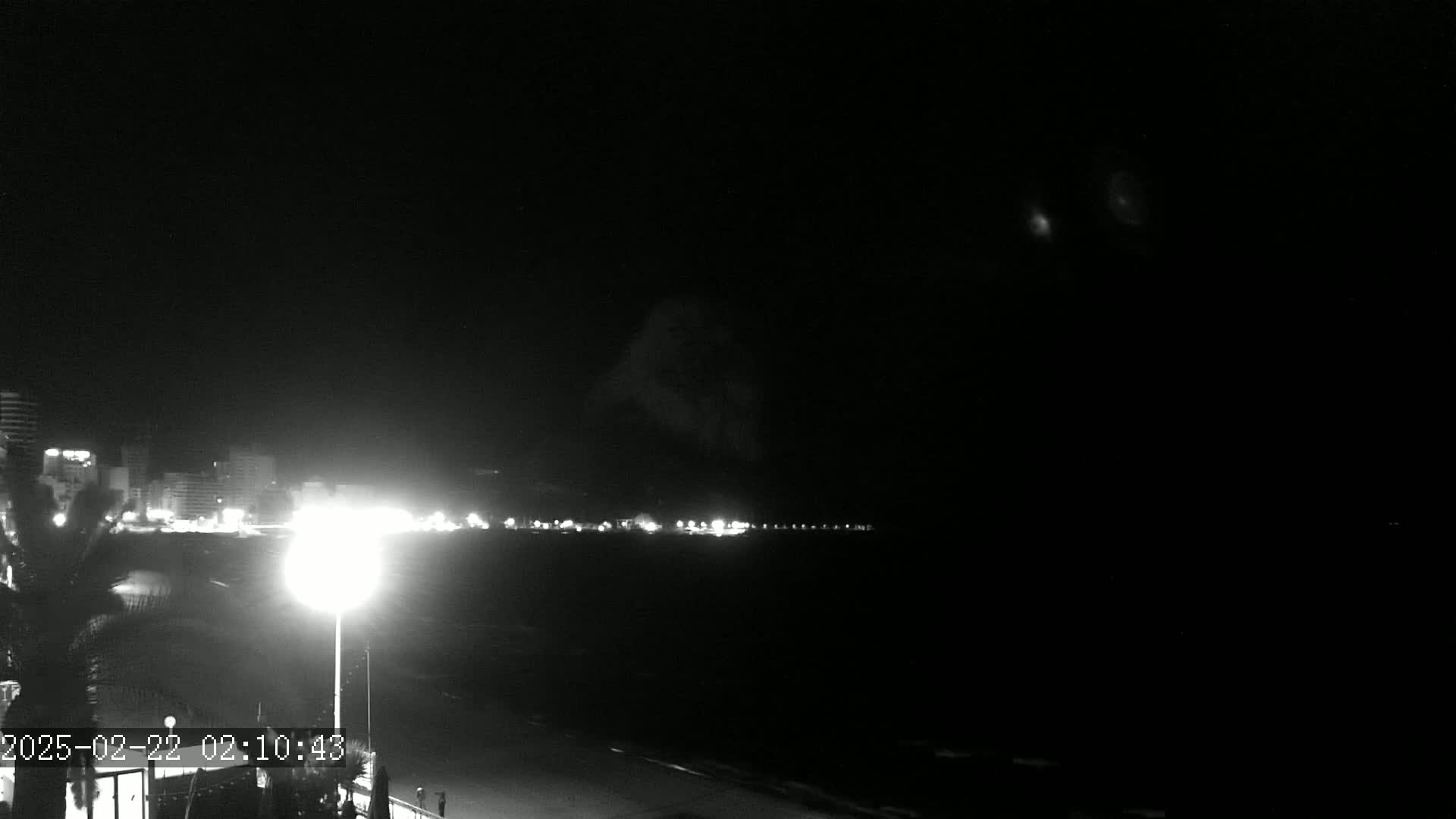 A nighttime, low-light view of a coastal city with faint lights illuminating the beachfront and a dark sky above.