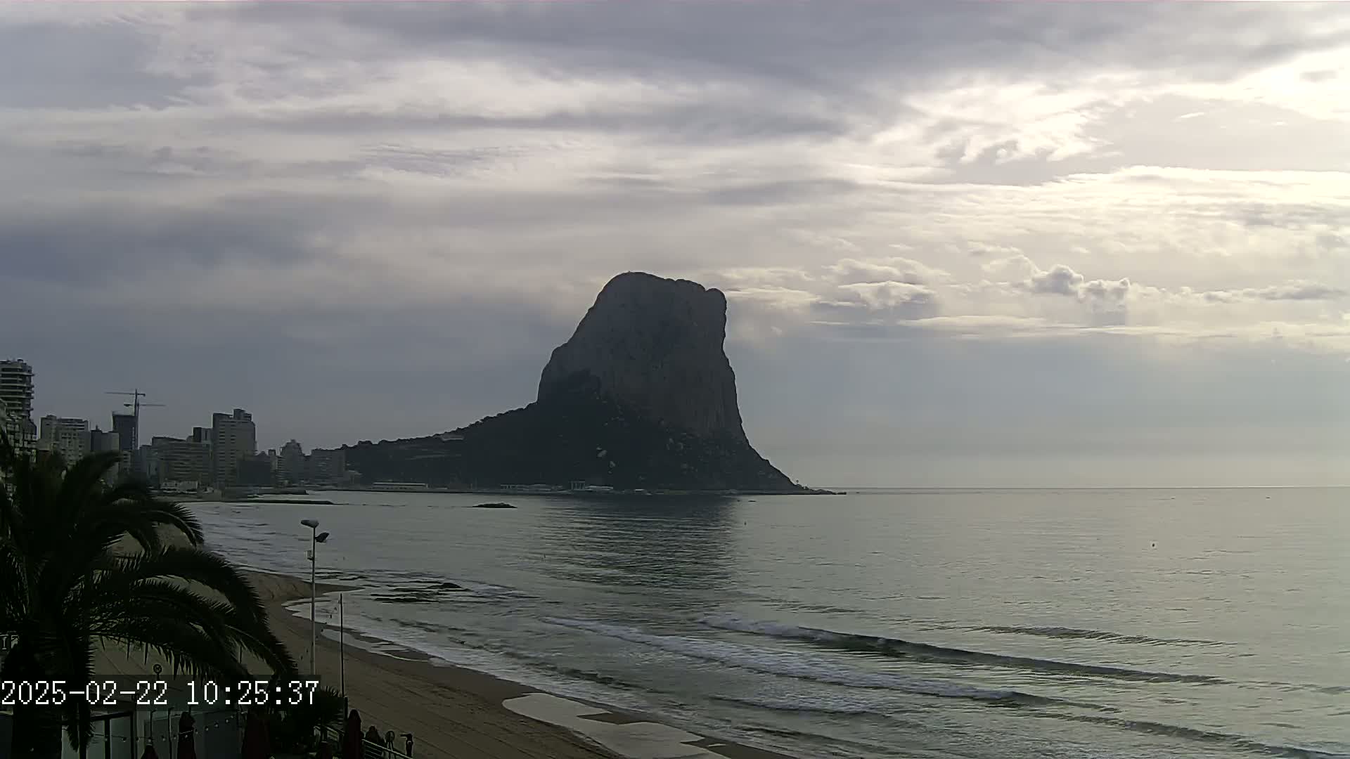 A large, dark rock formation rises from the sea under a cloudy sky, with a sandy beach and buildings visible in the foreground.