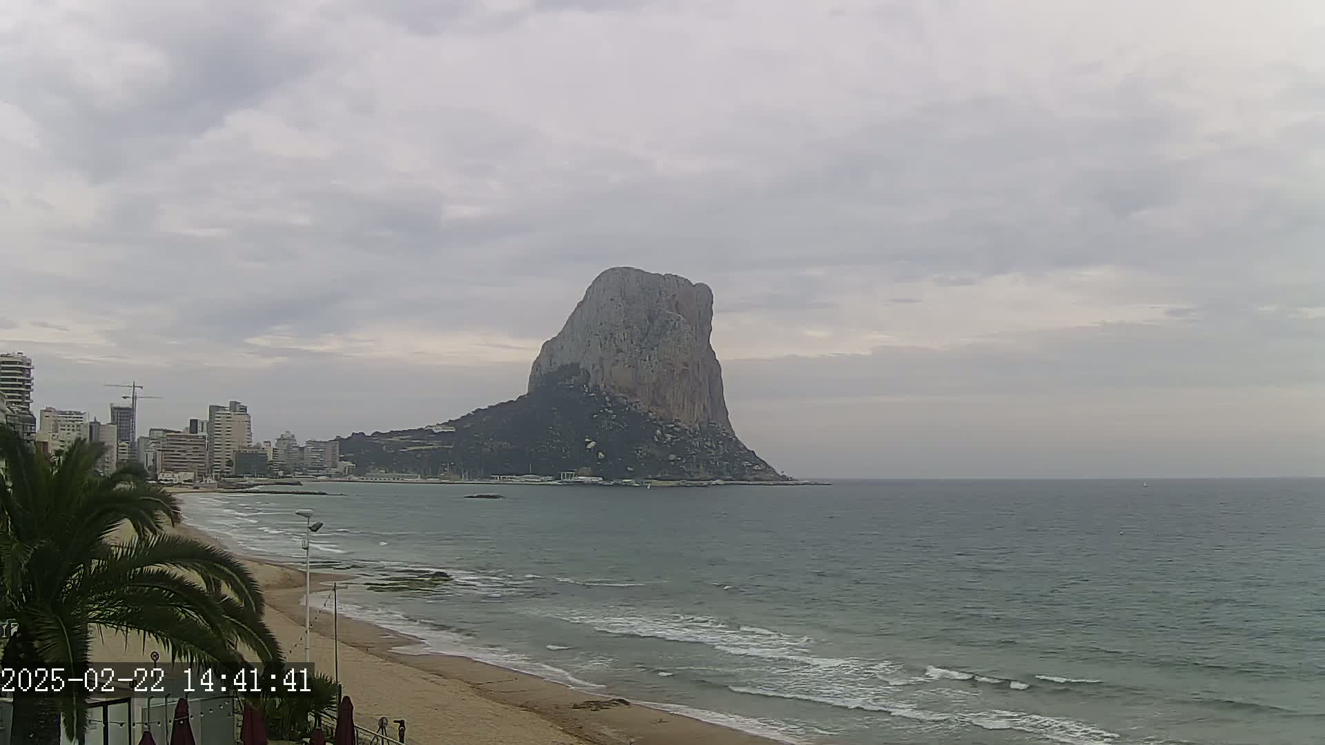 A large, rocky mountain rises from the sea under an overcast sky, overlooking a sandy beach and a cityscape.