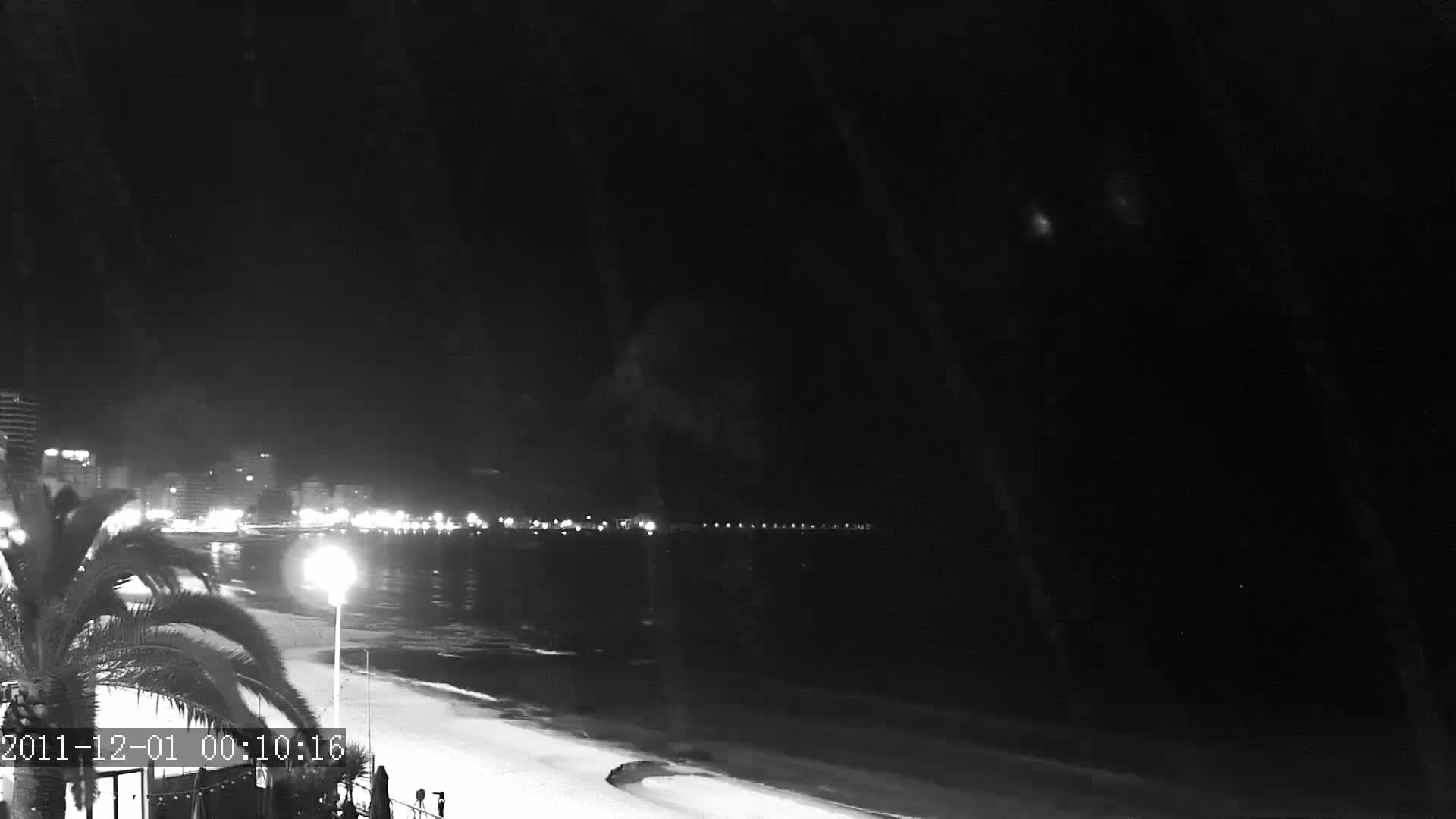 A nighttime, low-light view of a beach with a city skyline in the distance, showing a calm sea.