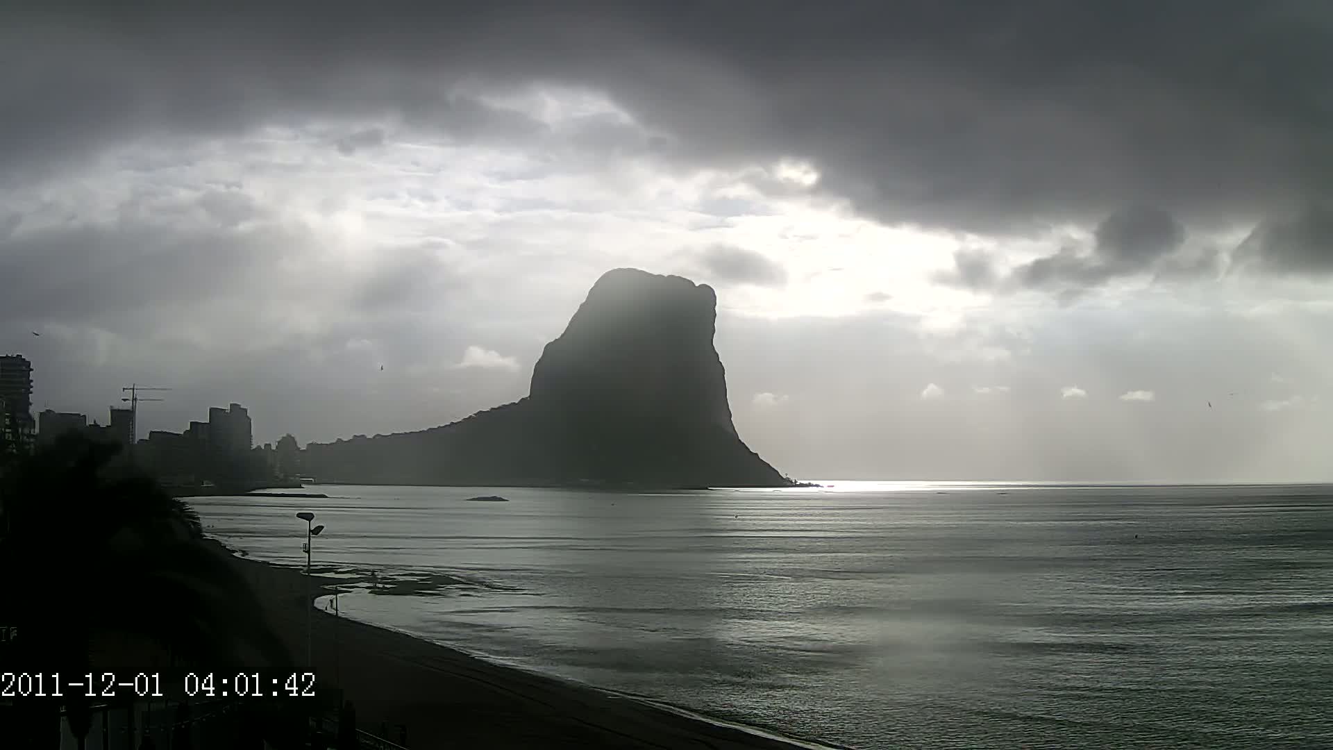 A large, dark mountain rises from the sea under a cloudy, overcast sky, with a beach and city buildings visible in the foreground.