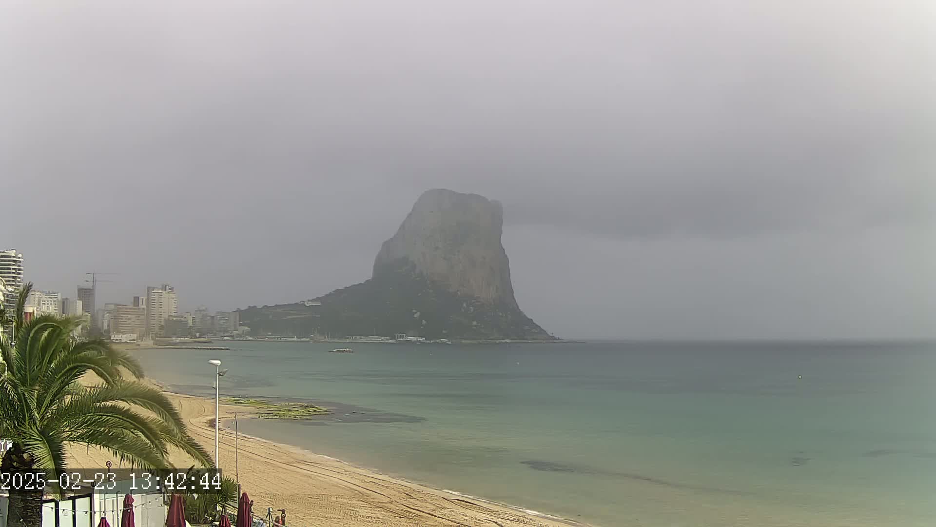 A large, rocky mountain rises from the sea on an overcast day, viewed from a sandy beach with buildings and palm trees in the foreground.