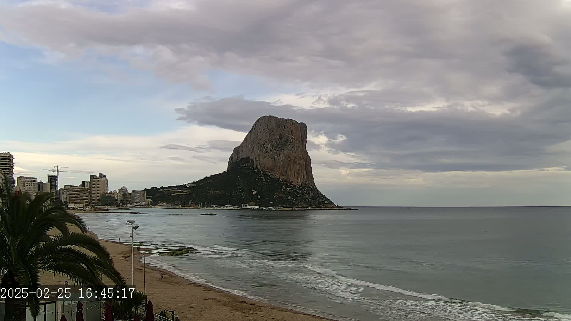 A large, rocky island sits in the ocean under a cloudy sky, with a sandy beach and buildings visible along the shoreline.