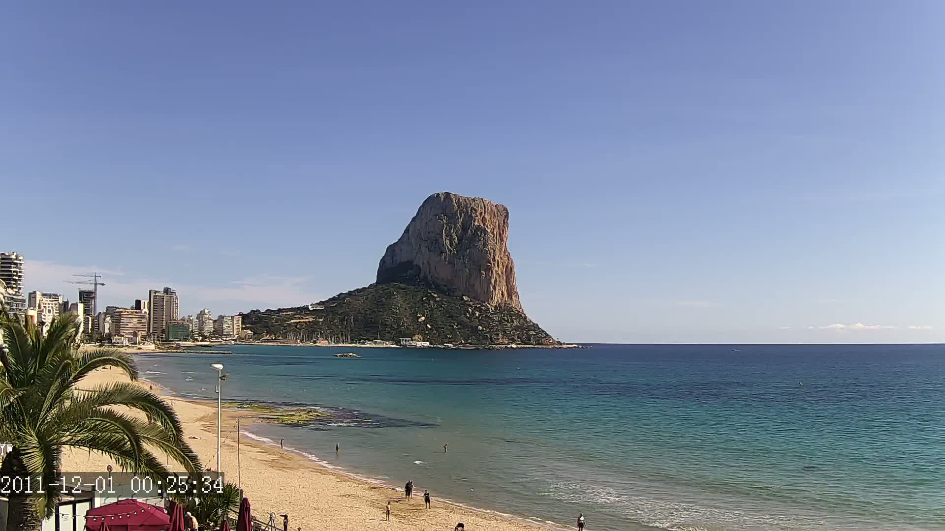 A sandy beach with a few people on it stretches out to a calm, teal-colored sea, beyond which a large, rocky, isolated mountain rises under a clear blue sky.