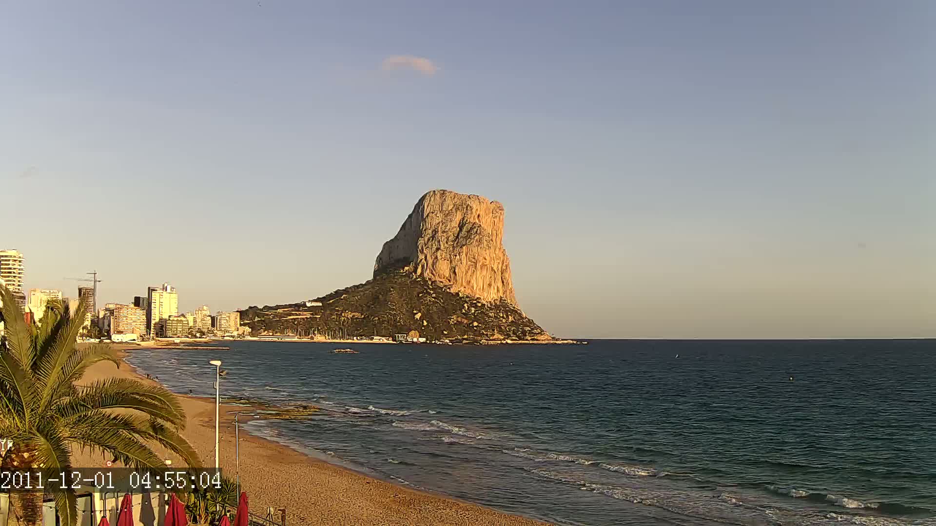 A large, rocky island stands in a calm sea under a clear, light-blue sky, near a sandy beach with a city skyline in the background.