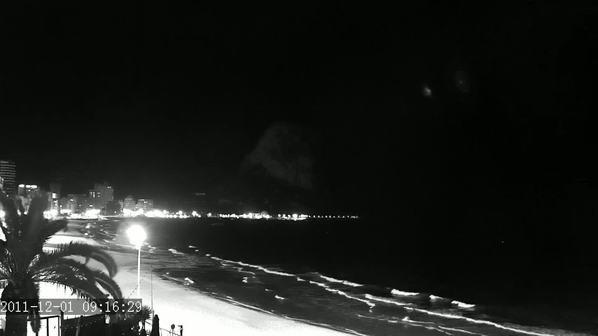 A nighttime, black and white view of a beach with gentle waves lapping the shore, illuminated by city lights in the distance and a single lamppost nearby.