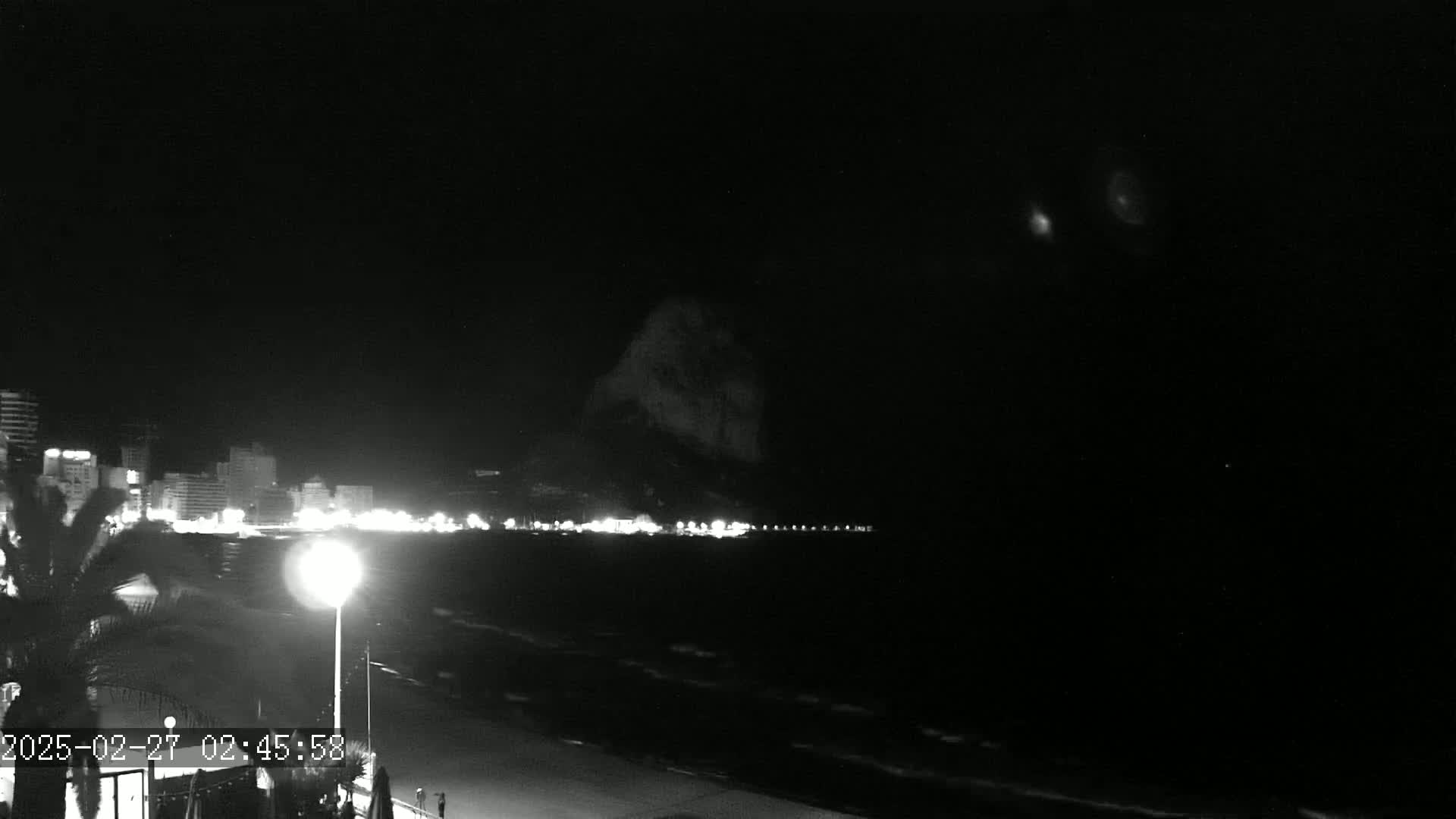 A nighttime beach scene shows a dark, wavy ocean, a line of lit buildings, and a large, dark hill or mountain in the background under a clear, dark sky.