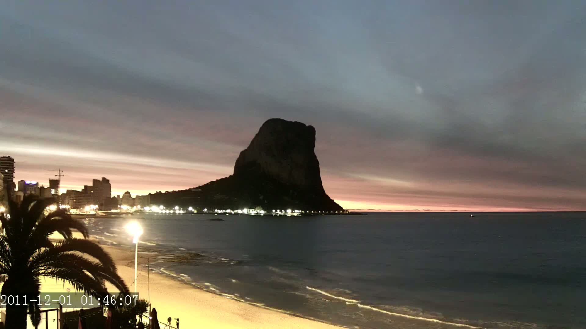 A large, dark rock formation stands silhouetted against a dusky, pink-tinged twilight sky over a beach with a city skyline and gently lapping waves.