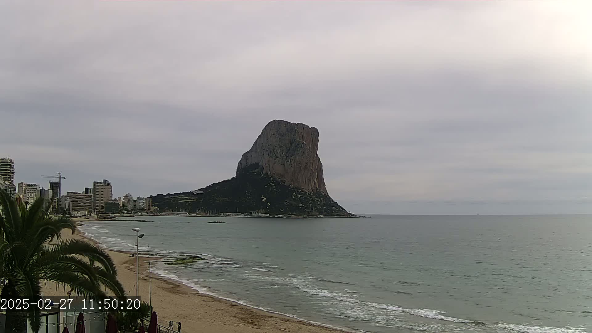 A large, dark rock formation rises from the sea near a sandy beach and city skyline under an overcast sky.