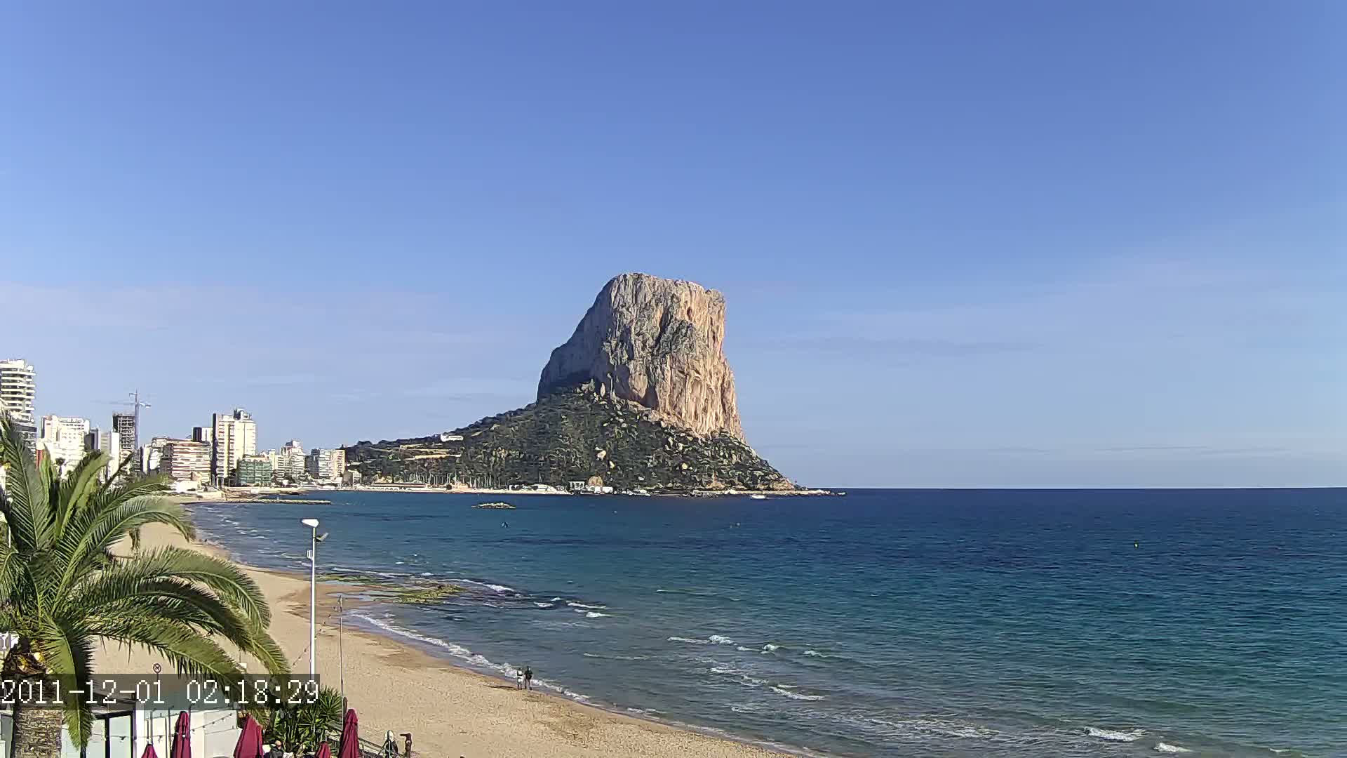 A sandy beach with calm ocean waves stretches towards a large, rocky cliff rising from the water, under a clear blue sky.