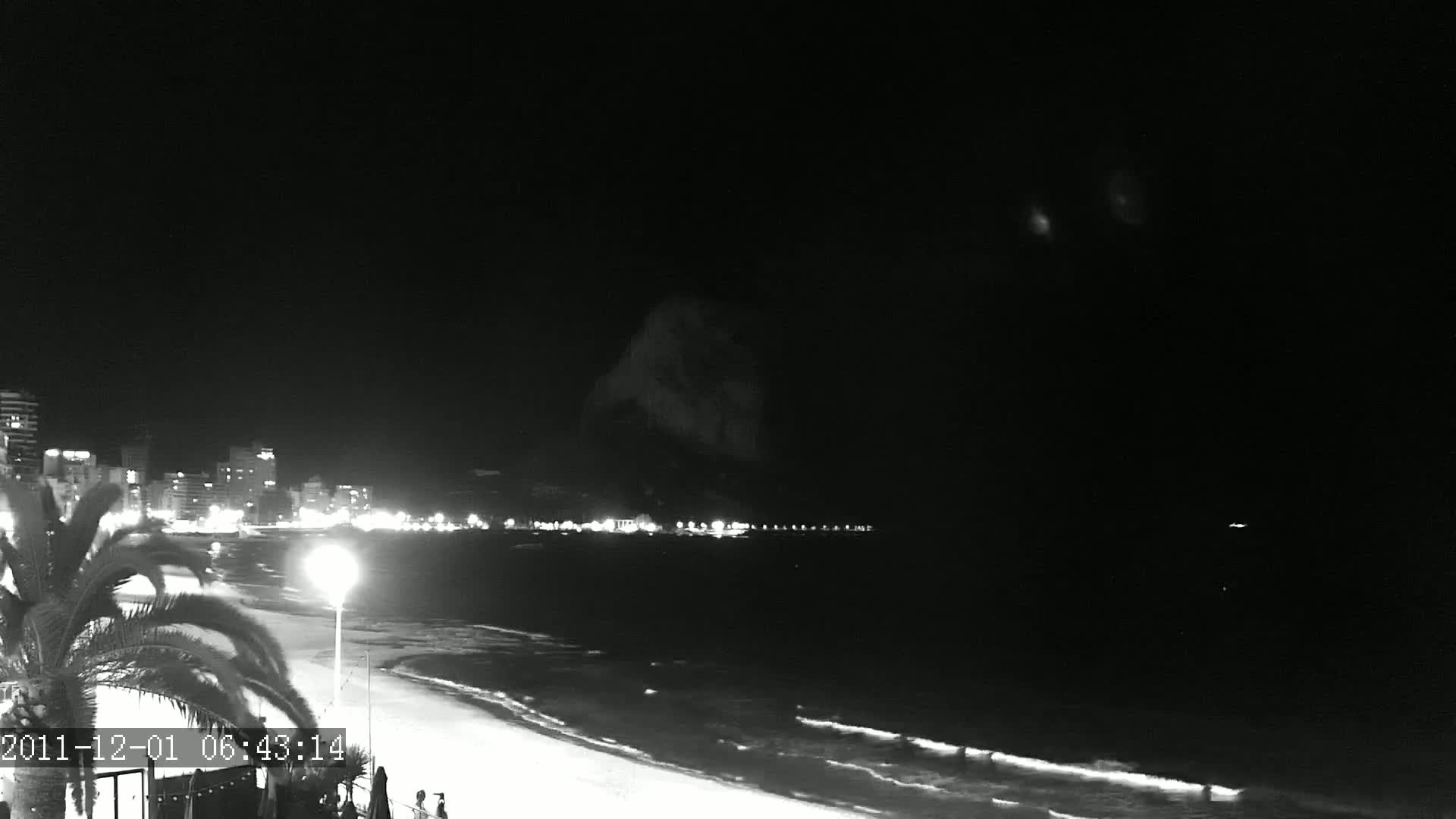 A nighttime beach scene shows a dark ocean with gentle waves lapping a sandy shore, illuminated by a city skyline across the water.
