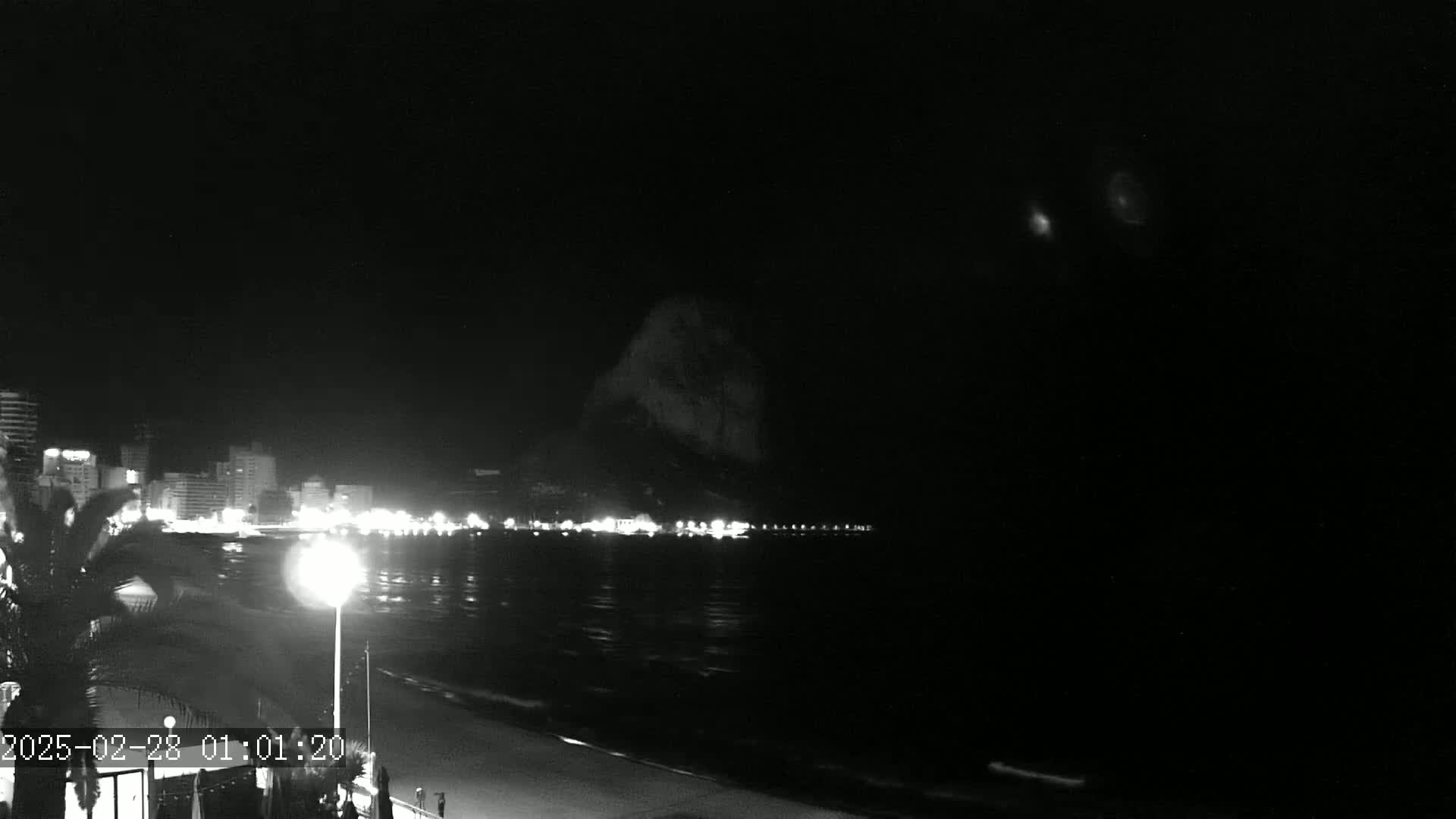 A nighttime beach scene shows a dark body of water reflecting the lights of a city skyline and a large, dark hill in the distance under a clear, dark sky.