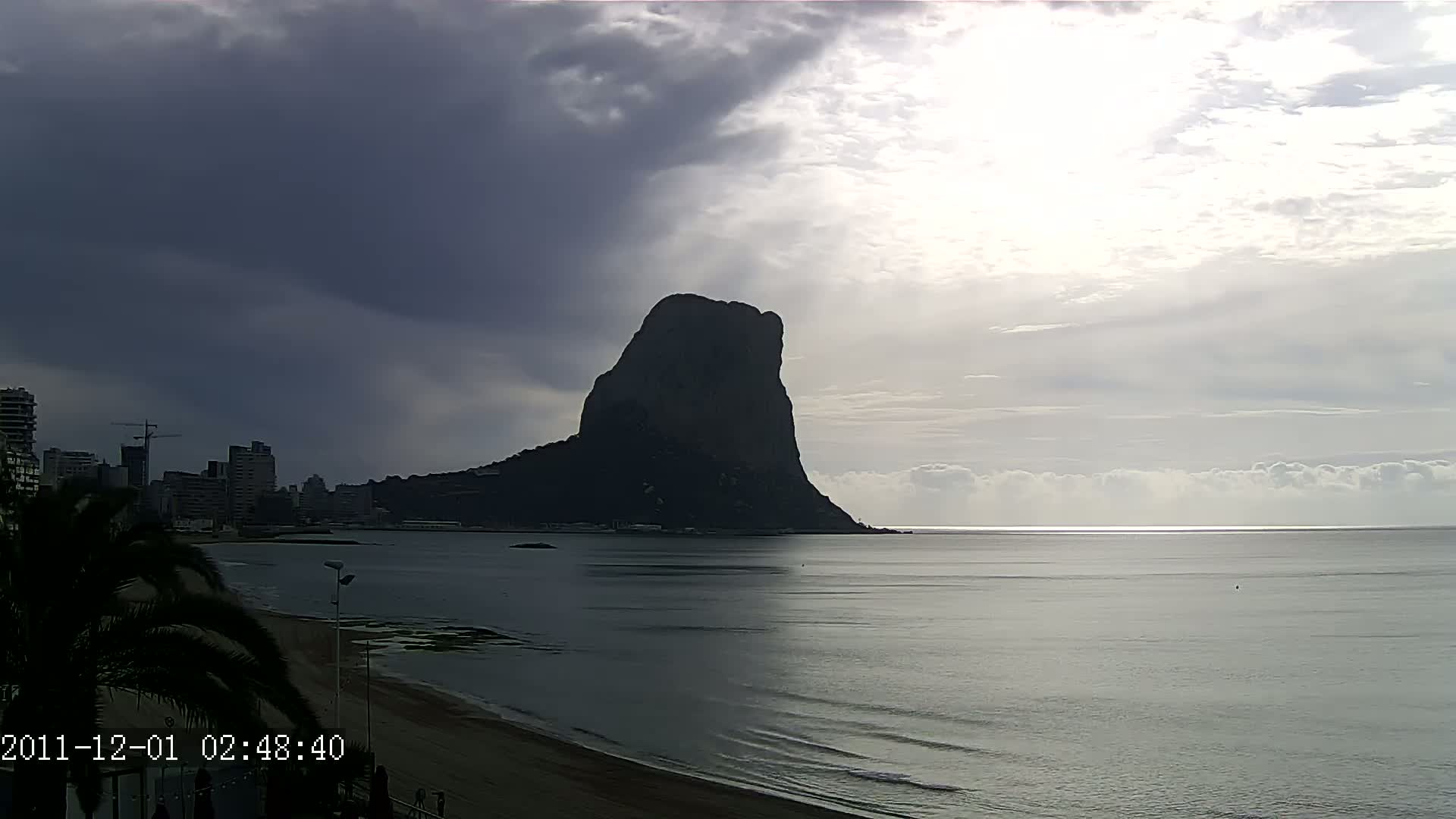 A large, dark mountain rises from the sea under a partly cloudy sky, with a city visible in the background on the left.