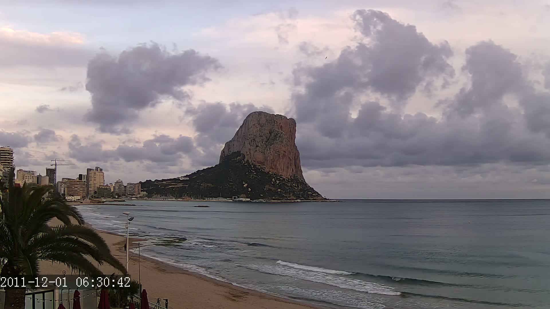A large, rocky mountain rises from the sea near a beach and city under a cloudy sky.