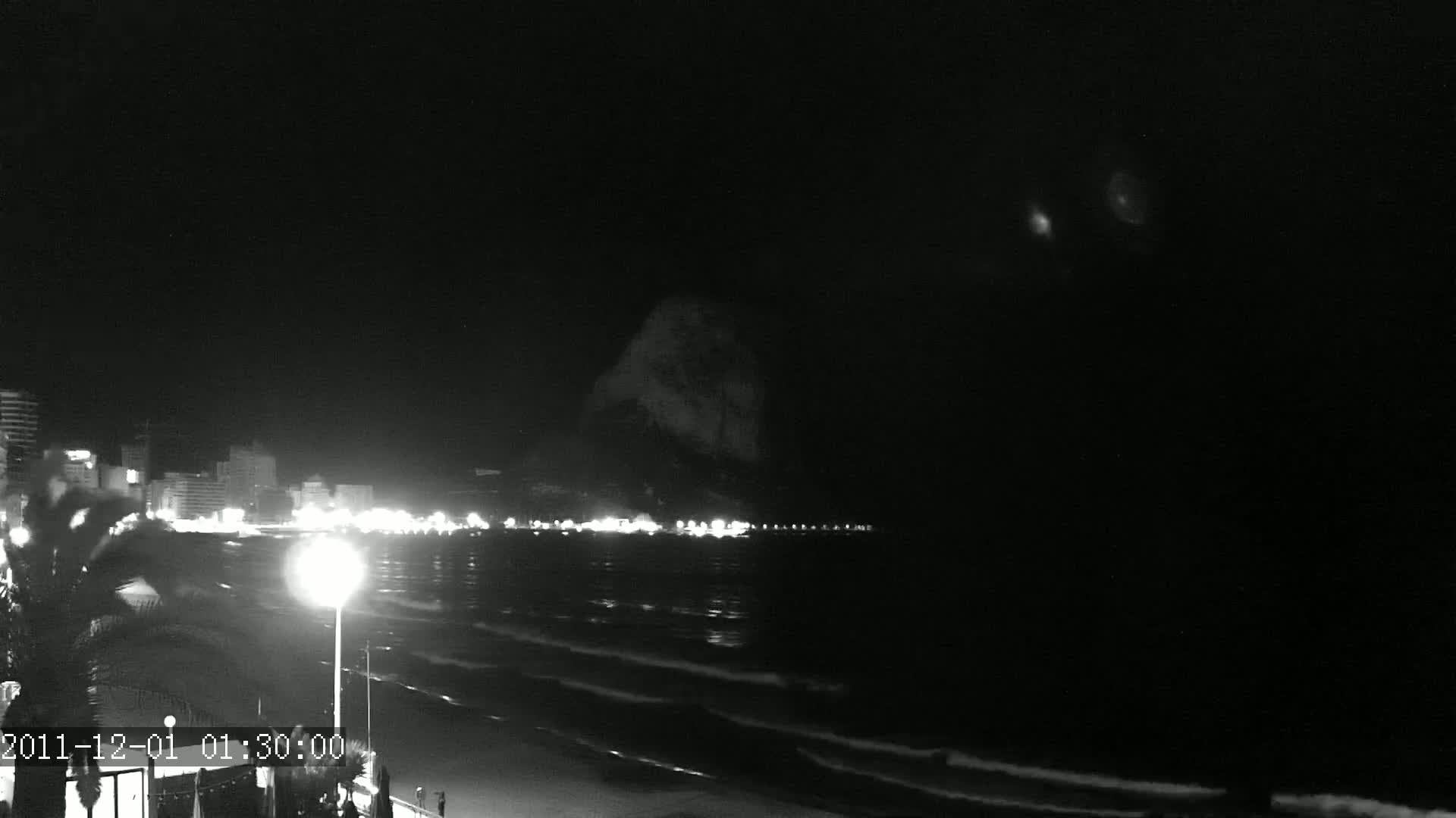 A nighttime, low-light view of a dark ocean with gentle waves lapping a shoreline, illuminated by city lights and a streetlamp, with a large, dark landmass visible in the distance.