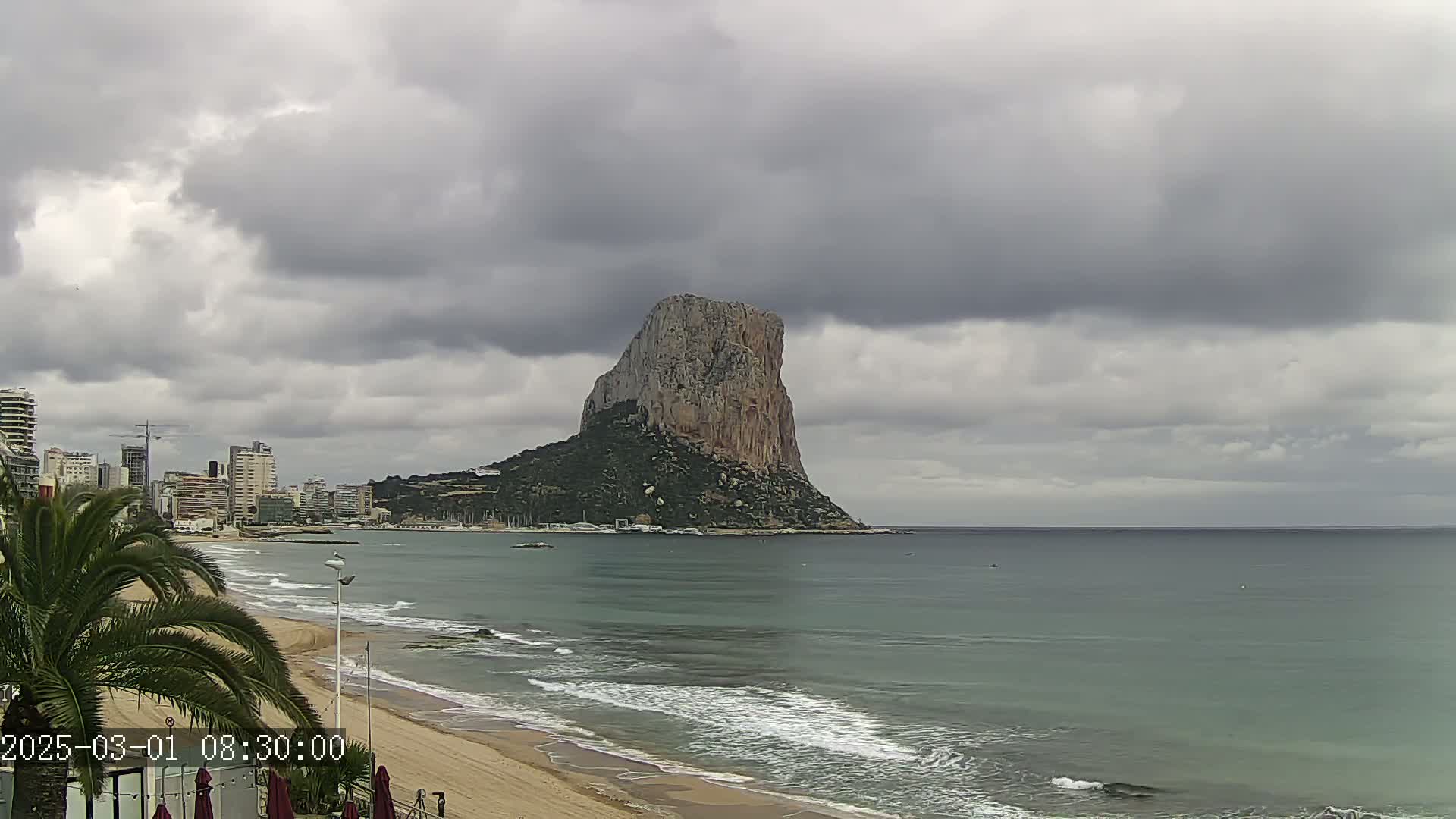 A large, rocky cliff stands prominently in the distance across a calm, grey sea under an overcast sky, with a sandy beach and a cityscape visible in the foreground.