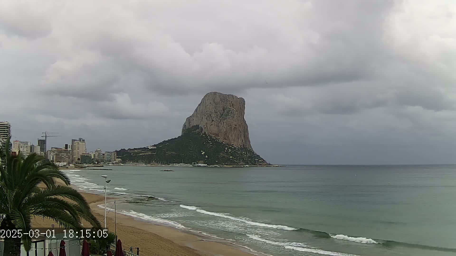 A large, rocky mountain rises from the sea under a cloudy sky, overlooking a sandy beach with gentle waves and a cityscape in the distance.