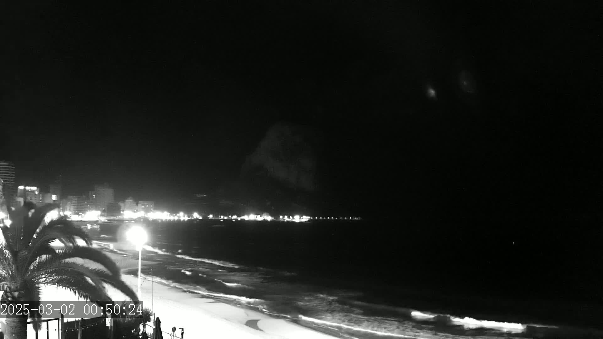 A nighttime beach scene shows a dark, mostly clear sky over a gently lapping ocean with a city's lights visible in the distance.