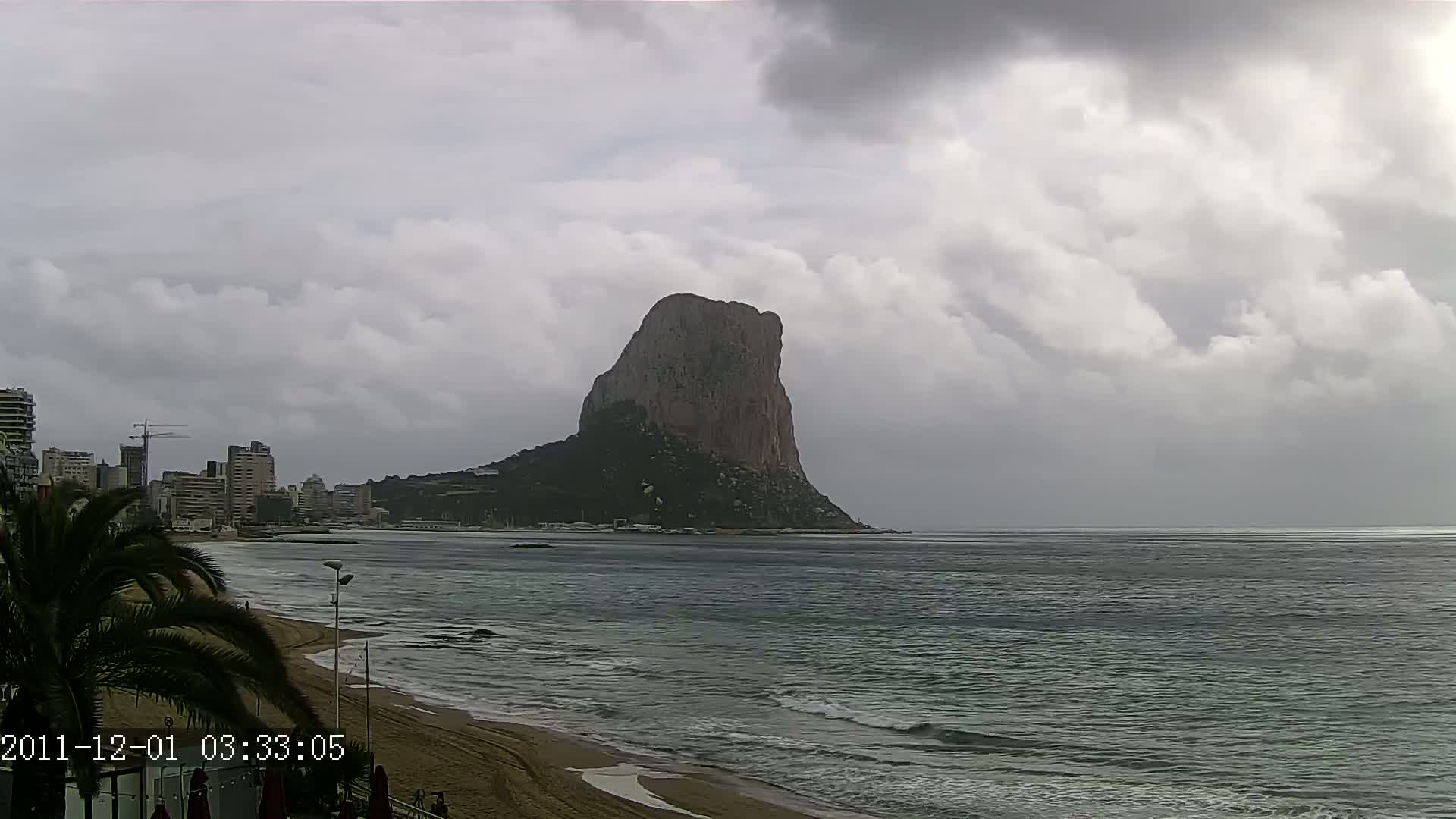 A large, dark rock formation rises from the sea under a cloudy sky, with a city and sandy beach visible in the foreground.