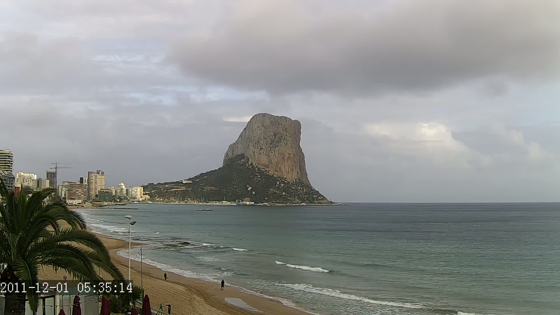 A large, rocky outcrop rises from the sea under a cloudy sky, overlooking a sandy beach and a cityscape.