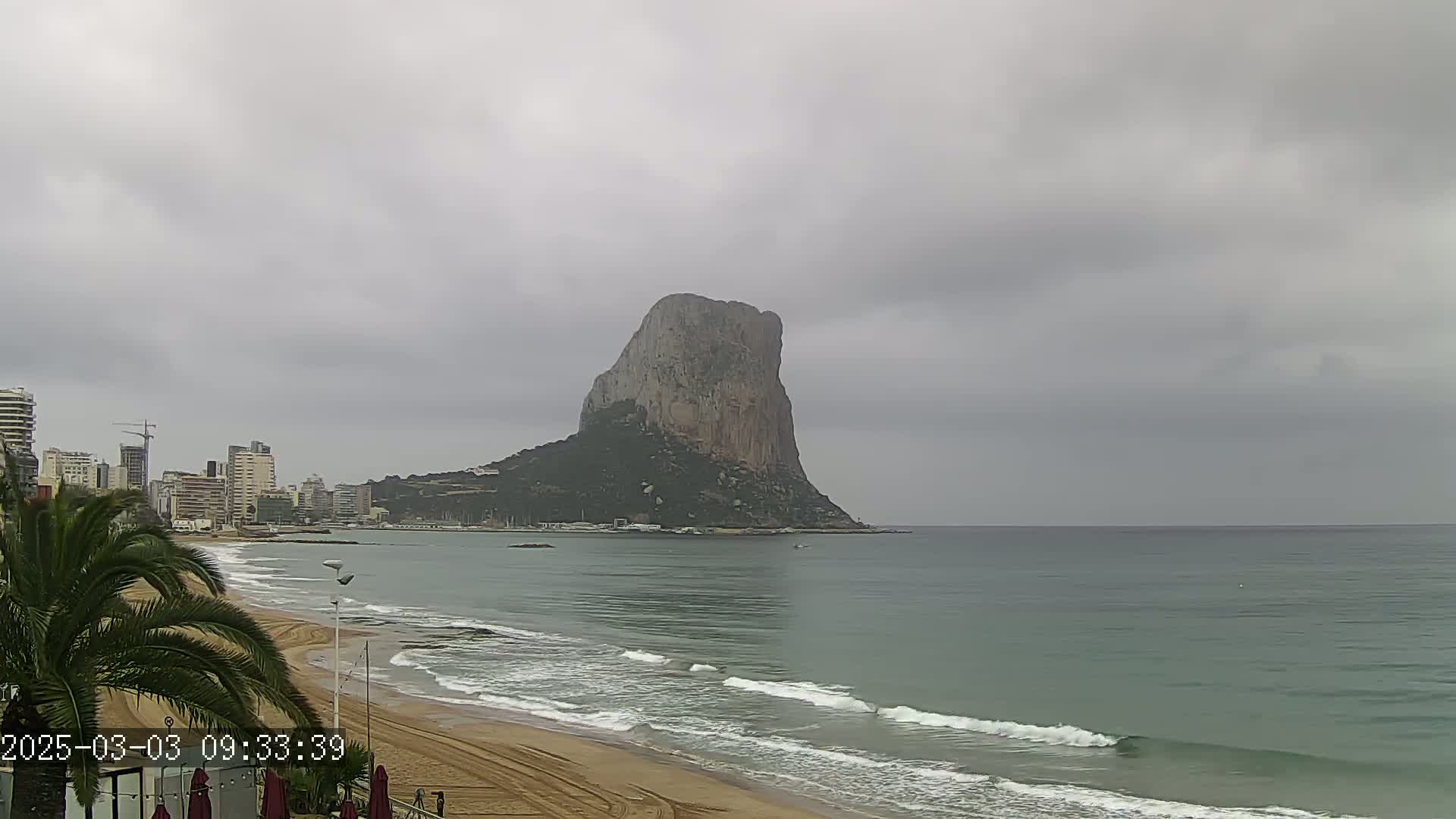A large, rocky mountain rises from the sea near a sandy beach and a line of buildings under an overcast sky.