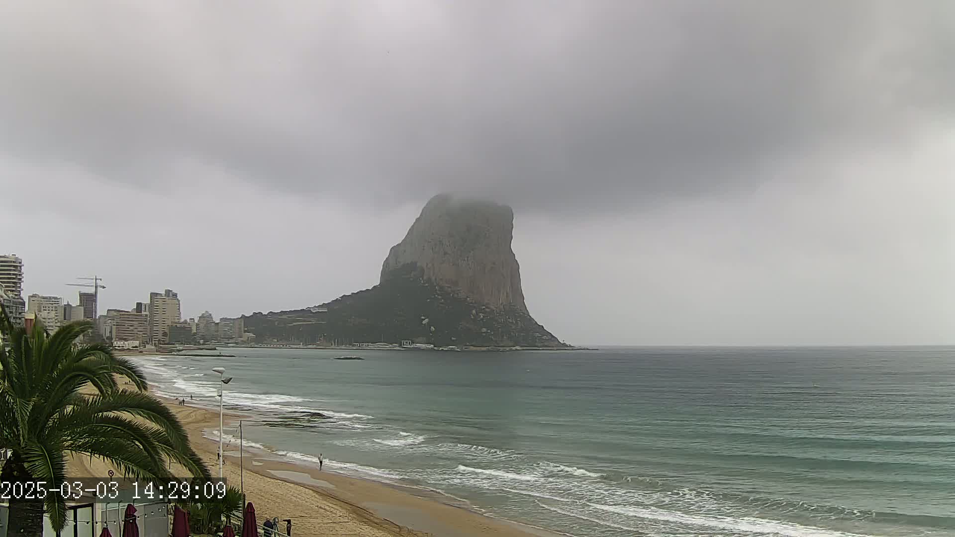 A large, dark rock formation is visible across a calm sea from a sandy beach under an overcast sky.