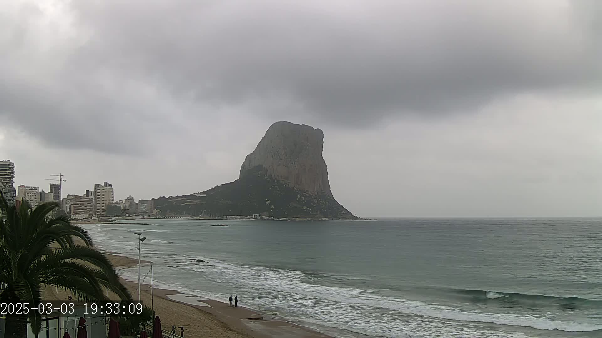 A large, dark-colored rock formation rises from the sea under an overcast sky, with a sandy beach and buildings visible in the foreground.
