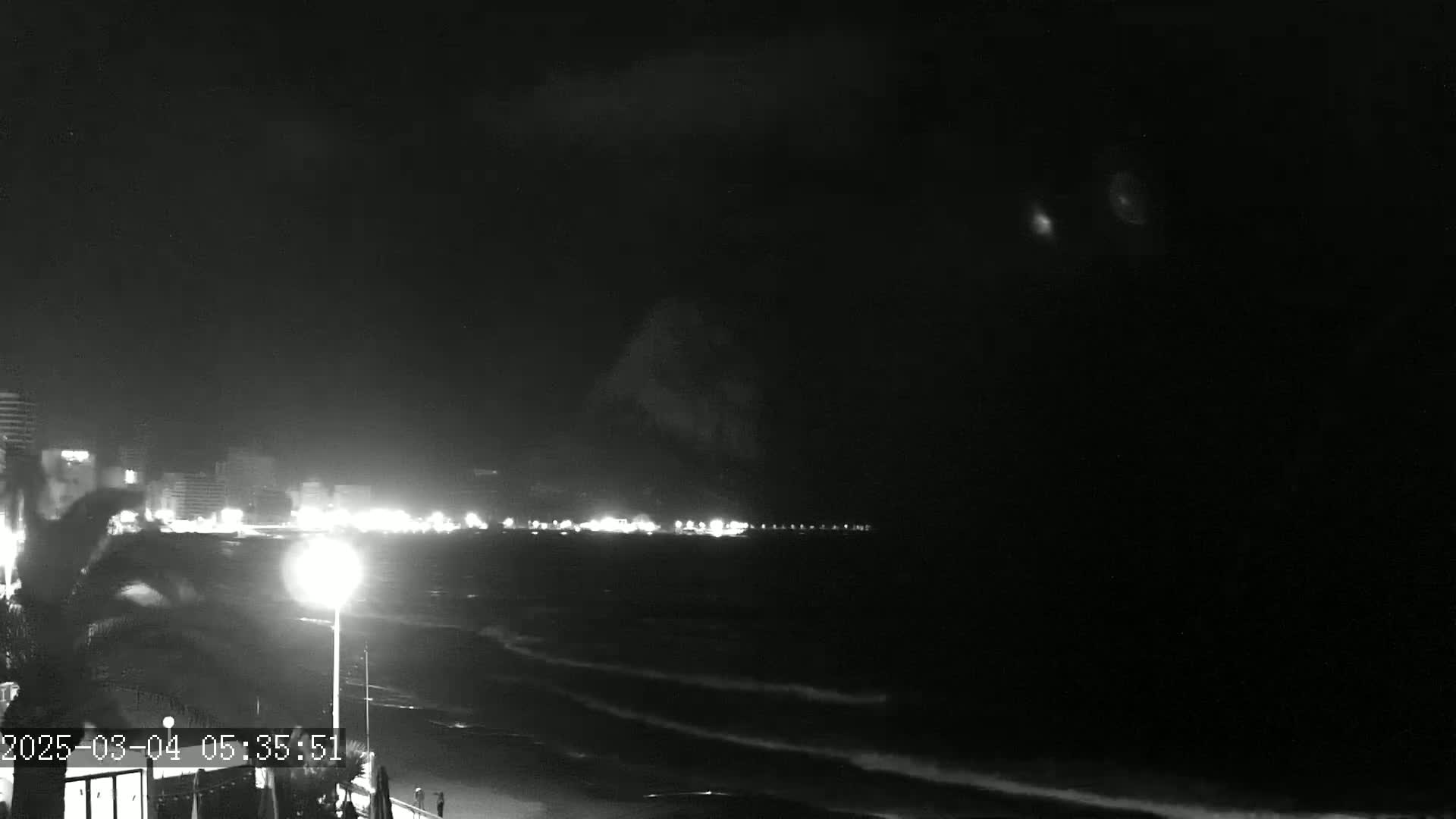 A nighttime, low-light view shows dark ocean waves lapping a shore, with a city's lights visible in the distance under a mostly clear, dark sky.