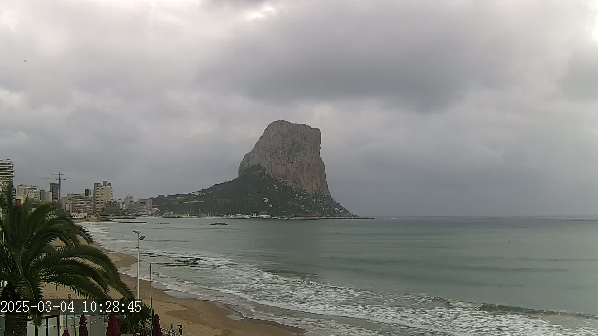 A large, rocky island rises from the sea under an overcast sky, with a sandy beach and city skyline visible in the foreground.