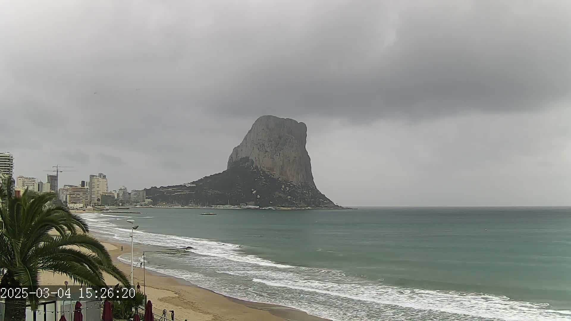 A large, rocky island sits in the distance across a calm, gray-green ocean, under an overcast sky; a sandy beach and a city skyline are visible in the foreground.