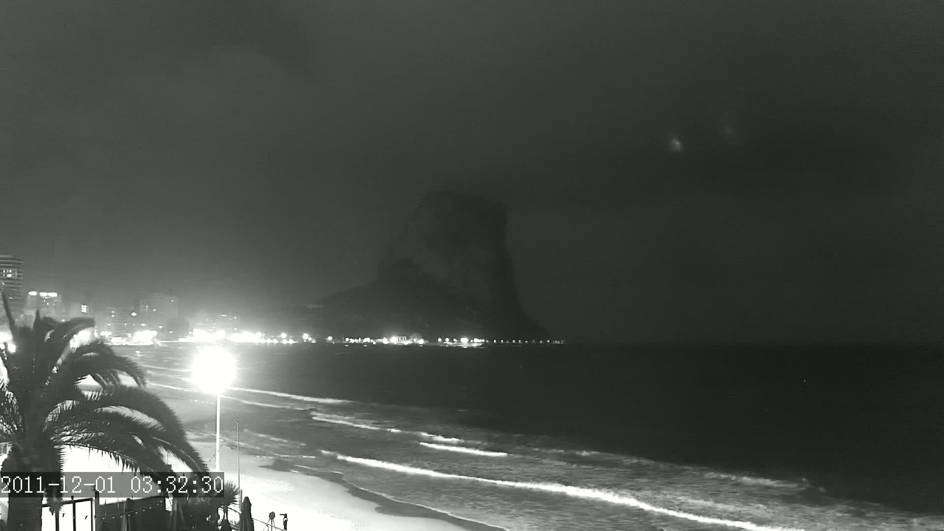 A nighttime beach scene shows gently rolling waves lapping at the shore, under a dark sky with a large, dark landmass visible in the distance.