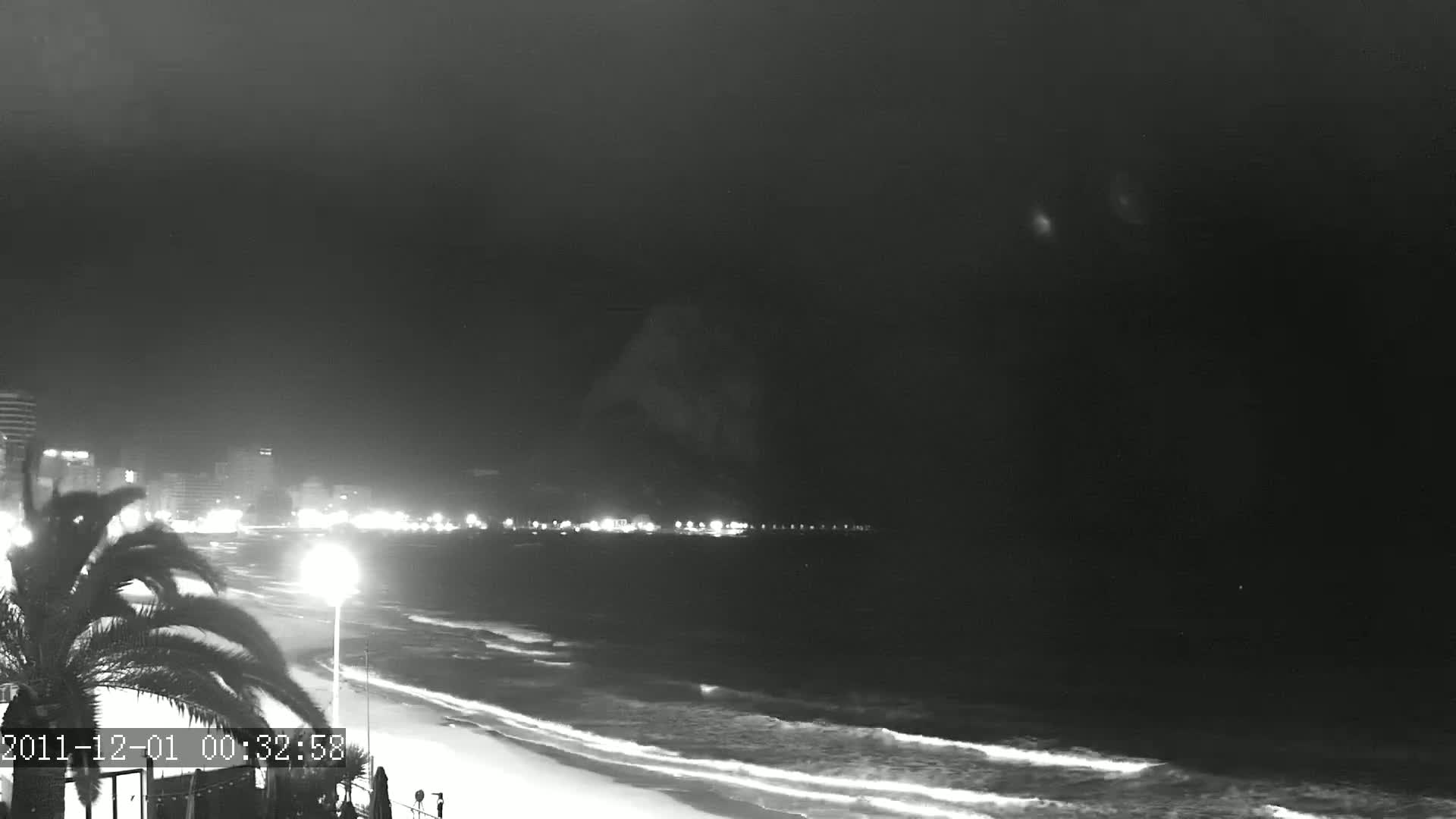 A nighttime beach scene shows a dark, overcast sky above gently lapping waves on a sandy shore, with city lights illuminating a distant beachfront.