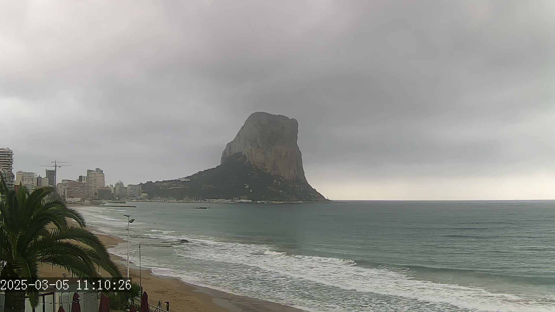 A large, imposing rock formation rises from the sea under a cloudy sky, as waves lap a sandy beach in front of a cityscape.