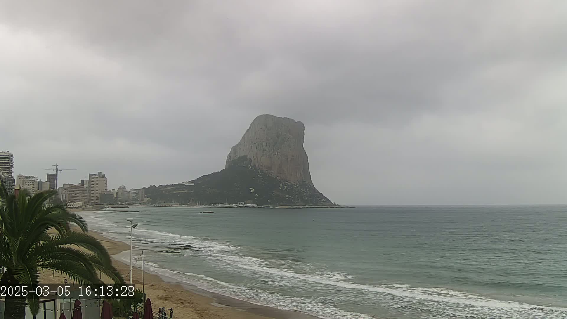 A large, rocky peninsula stands in the distance across a wavy sea under an overcast sky, with a sandy beach and a city skyline visible closer to the foreground.