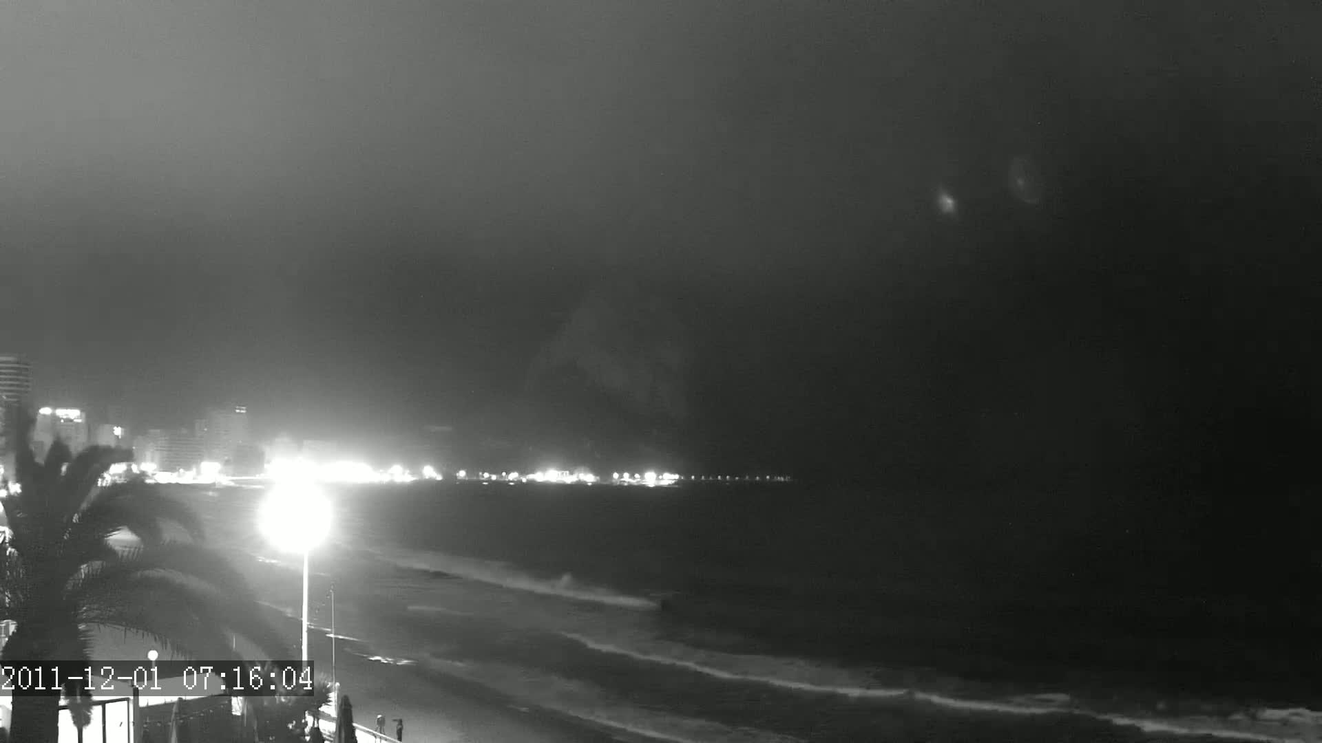 A nighttime beach scene shows gently rolling waves lapping the shore under a dark, overcast sky.