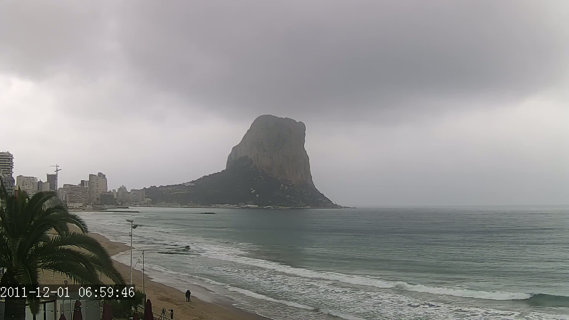 A large, imposing rock formation rises from the sea under a cloudy, overcast sky, with a sandy beach and buildings visible in the foreground.