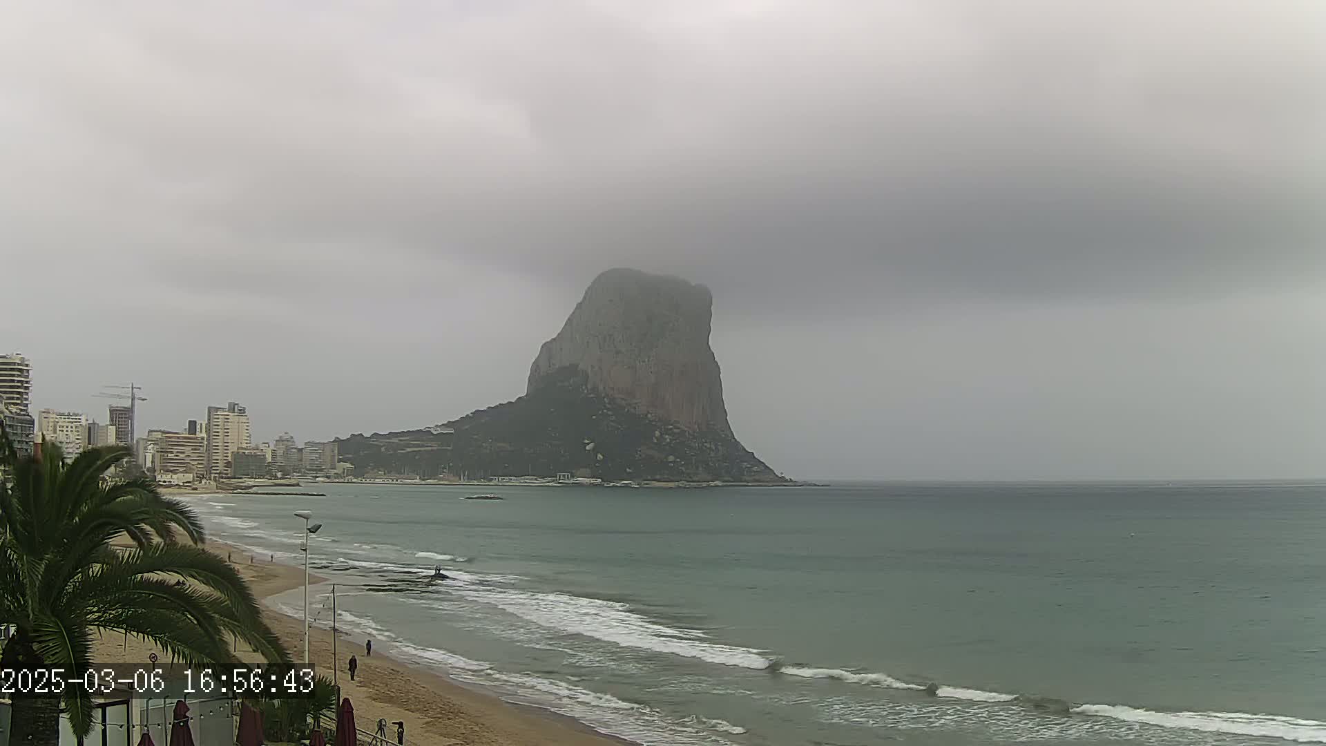 A large, rocky mountain rises from the sea under an overcast sky, with a sandy beach and buildings visible in the foreground.