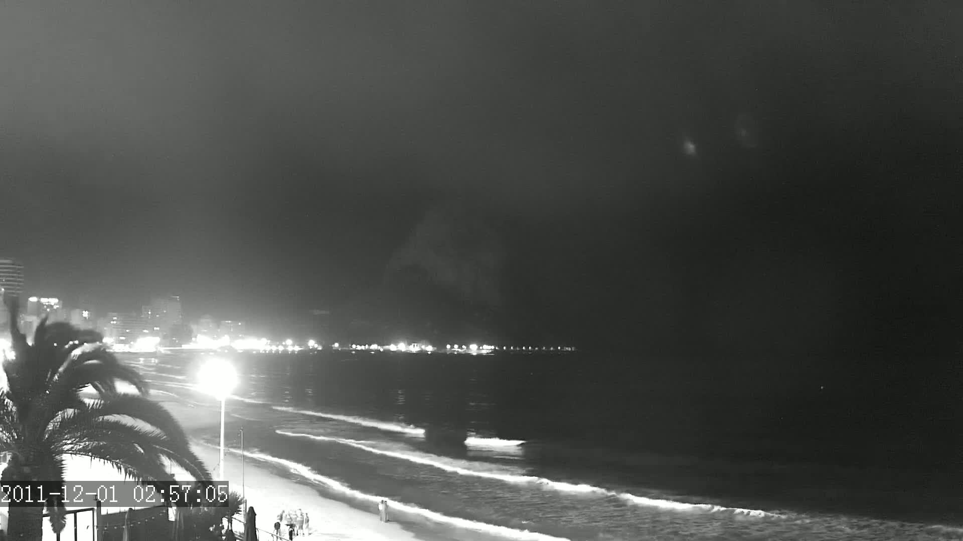 A nighttime beach scene shows gently lapping waves on a sandy shore, with a city skyline faintly illuminated in the distance under a dark sky.