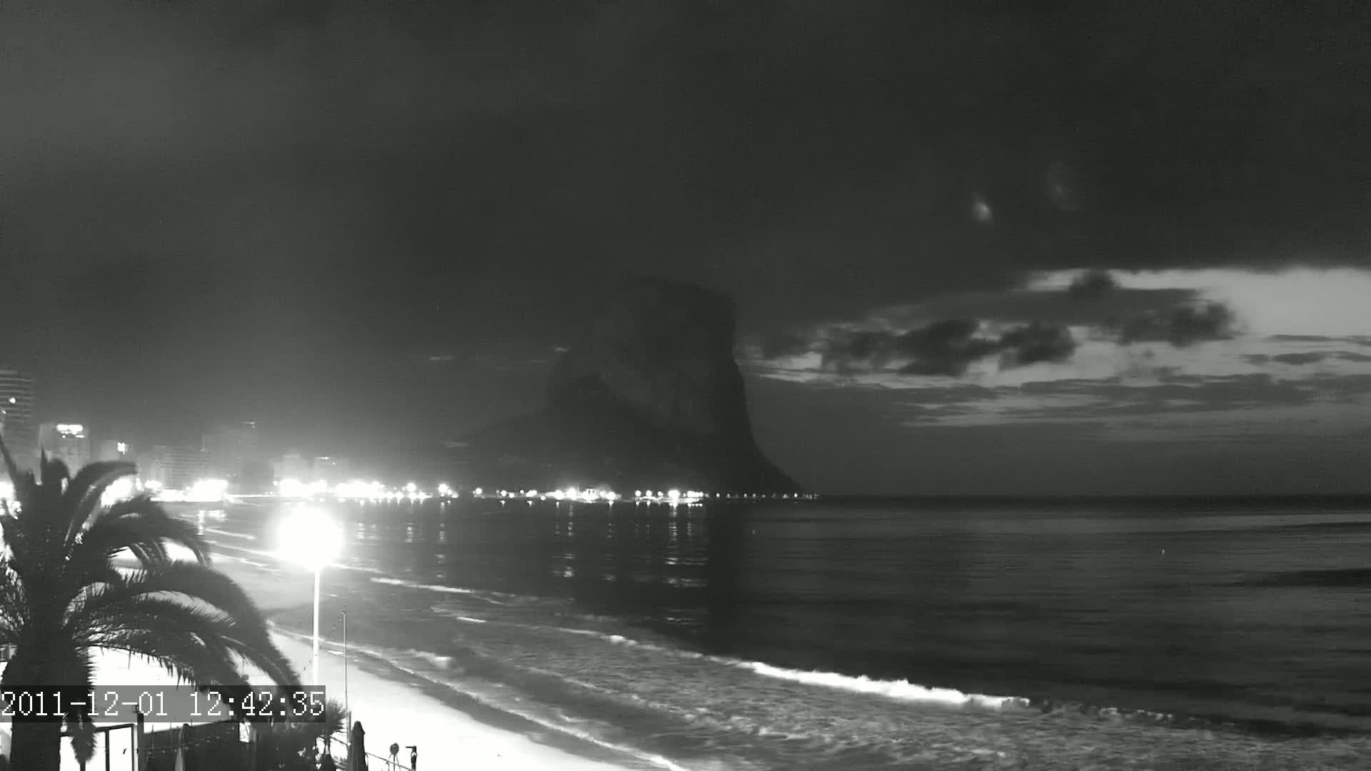A nighttime beach scene shows a dark, cloudy sky over a calm ocean with gentle waves lapping the shore,  illuminated by city lights and a large, dark rock formation in the distance.