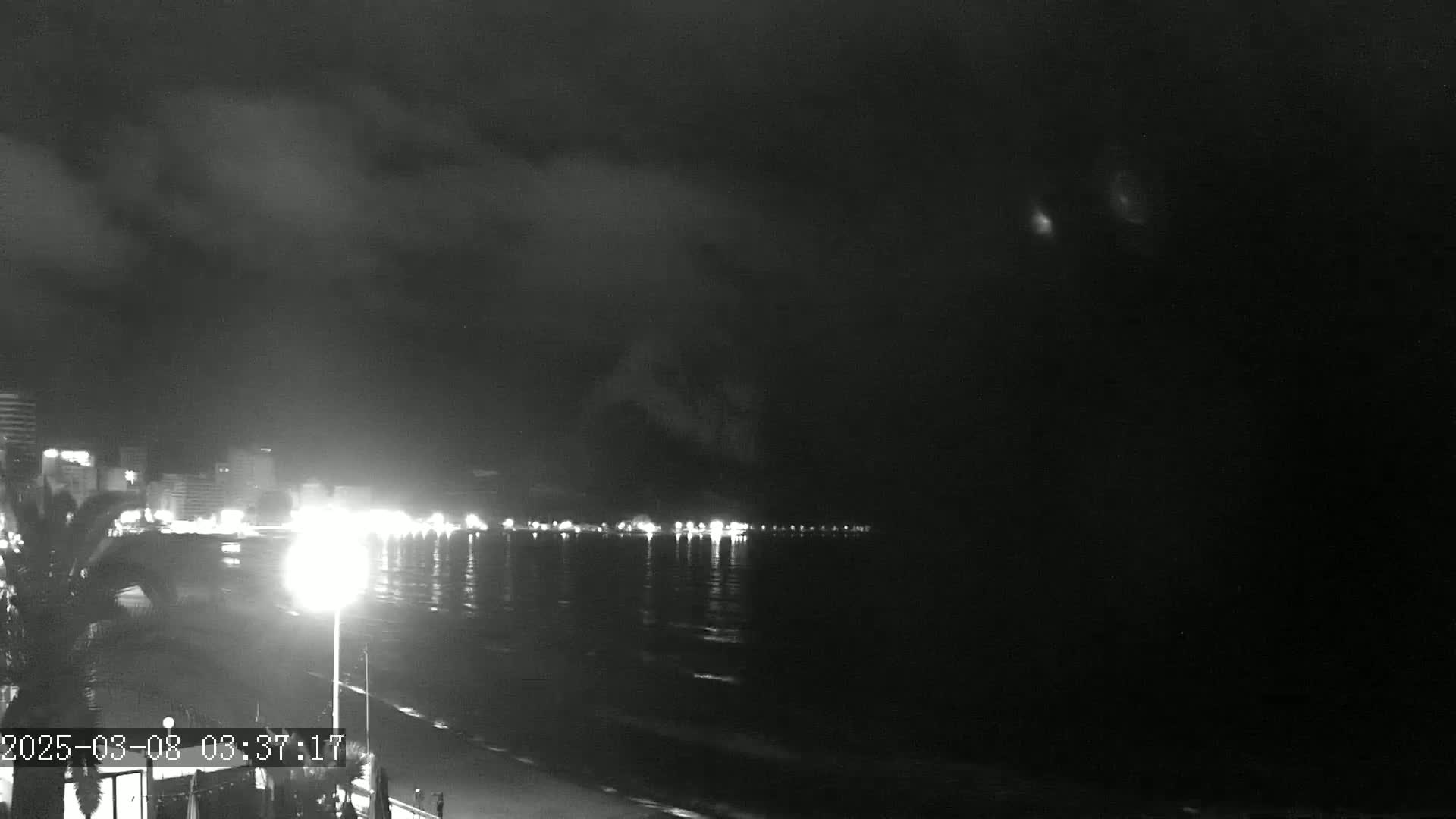 A nighttime view of a dimly lit beachfront, with buildings and lights visible across the water under a cloudy sky.