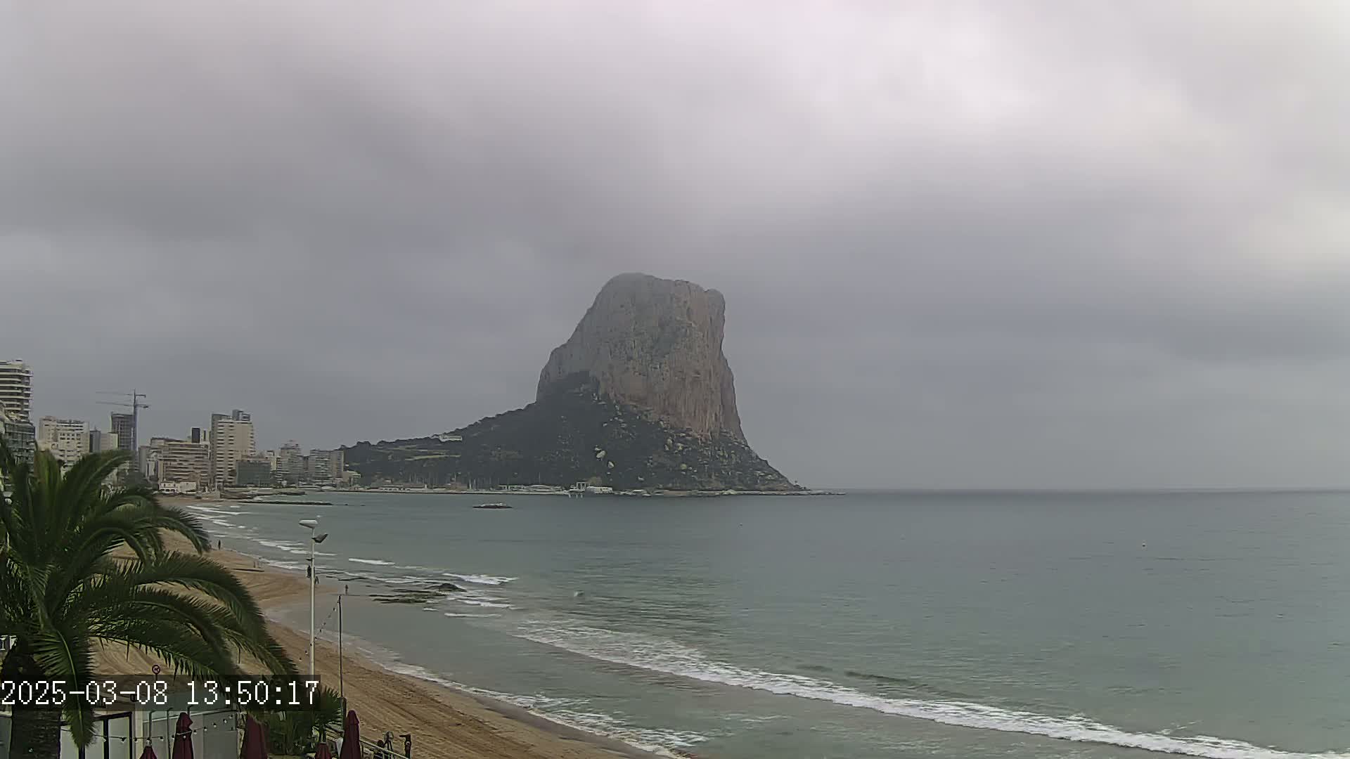 A large, rocky cliff stands in the distance overlooking a calm sea and sandy beach, under an overcast sky.